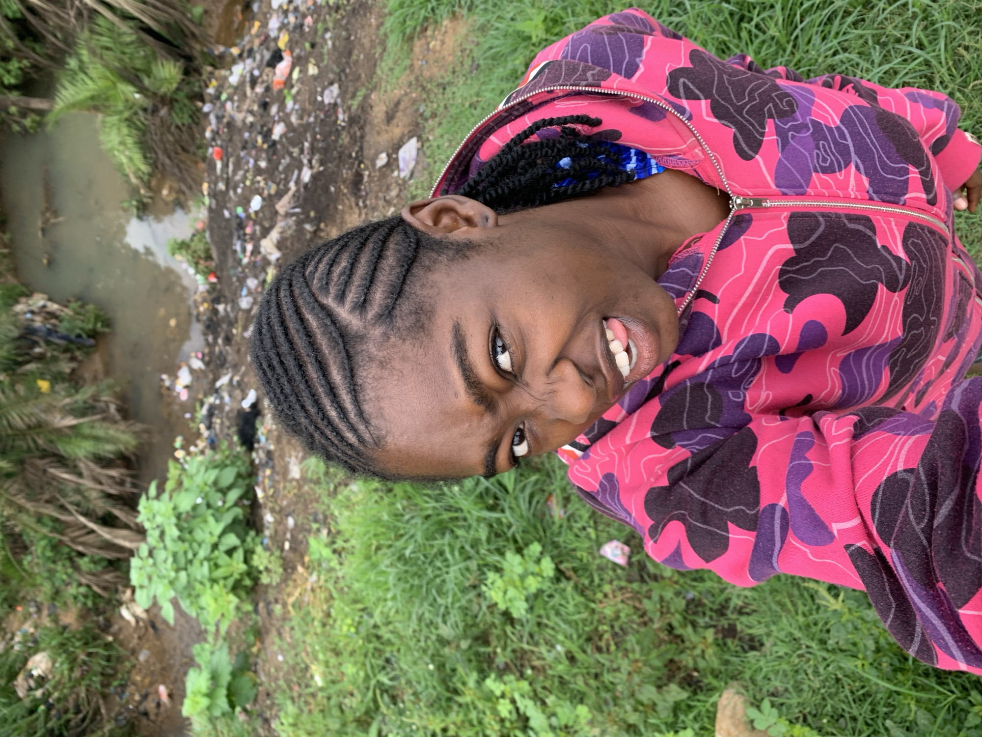 The image portrays an individual in Rafin Par, Nigeria, captured in a close-up, upward-angled shot. The subject, with dark skin, exhibits a playful or exaggerated facial expression, characterized by furrowed eyebrows, a wrinkled nose, and a visible tongue. Their hair is styled in black cornrow braids, with longer sections incorporating blue accents visible near the right ear. The individual wears a pink, purple, and black camouflage-patterned hooded sweatshirt with a prominent silver zipper.
The background transitions from lush green grass in the immediate foreground to a section of exposed dirt and mud. This area is heavily littered with various forms of refuse, including plastic containers and wrappers. Further into the background, a narrow, shallow, and murky water body, consistent with a stream or ditch, flows. The banks of this water feature are lined with green vegetation, including broad-leafed plants and several palm fronds, intermingled with additional scattered debris and waste materials.