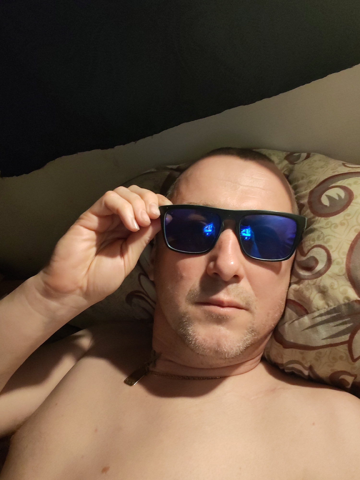 The image shows a man lying down indoors, likely on a bed or couch, with his head resting on a patterned pillow. He appears to be shirtless, revealing his upper chest and shoulders. He has fair skin, short hair with a receding hairline, and light stubble on his face. The man is wearing black-framed sunglasses with highly reflective blue lenses, which he is holding with his right hand, seemingly in the process of adjusting or removing them. A thin, gold-colored chain with a small, rectangular pendant hangs around his neck. He looks directly at the camera with a neutral expression. The background includes a dark, plain fabric in the upper left, a lighter-colored surface (possibly a wall or sheet) in the upper middle, and the patterned pillow to the right of his head. The overall lighting is somewhat dim, characteristic of an indoor setting with artificial light. There is no visible text. The scene is set in Bol'shie Mkhi, Russia.
