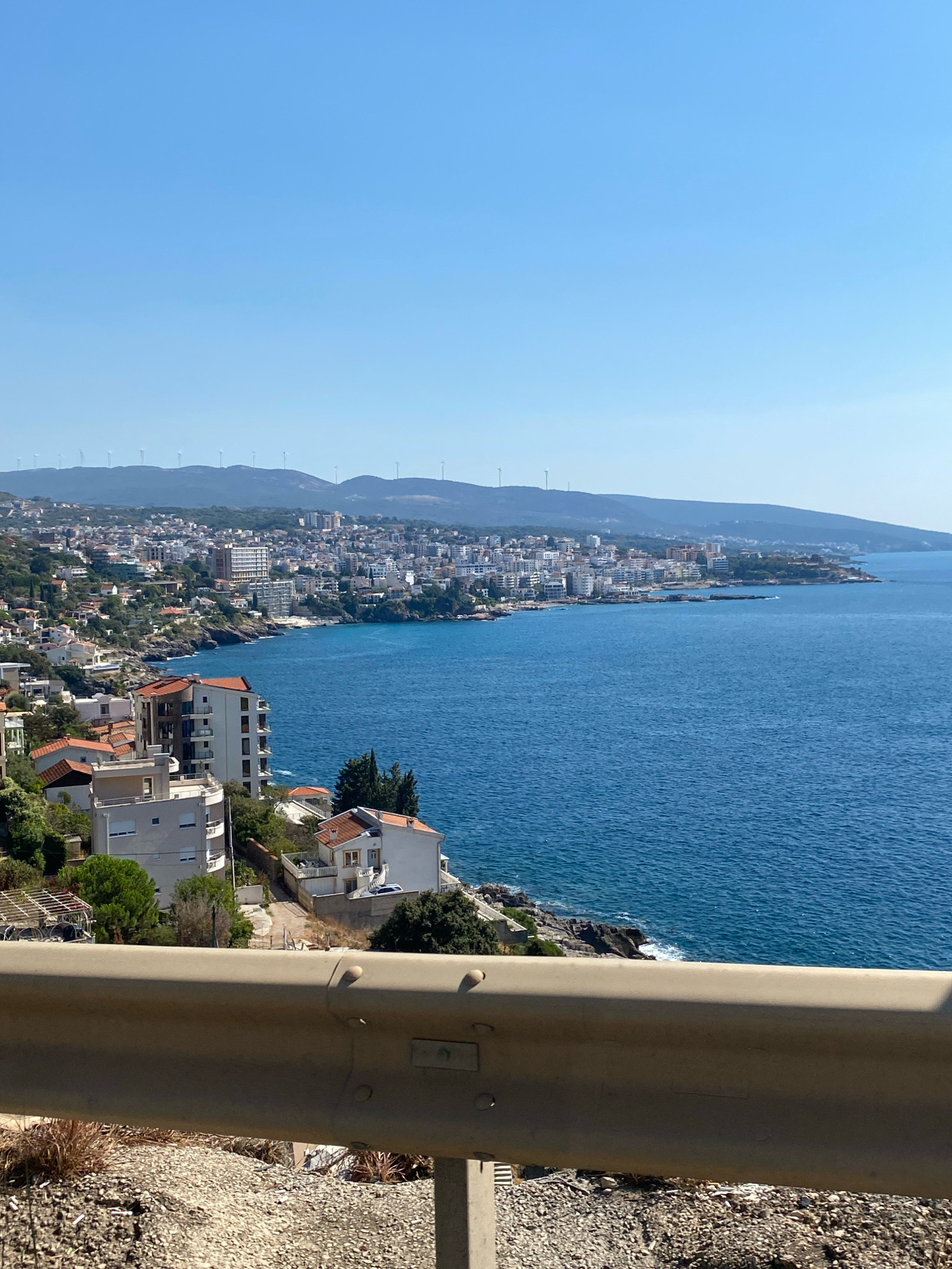 The image captures a panoramic view of the coastal city of Dobra Voda, Montenegro, under a clear blue sky. In the foreground, a beige guardrail runs horizontally across the lower portion of the frame, with rocky terrain and dry vegetation visible beneath it. Beyond the guardrail, the scene opens up to a densely populated coastline dotted with white and orange-roofed buildings nestled into a verdant hillside. The buildings range from smaller, residential structures to larger, multi-story apartment complexes and hotels, many of which have balconies overlooking the sea. The deep blue Adriatic Sea occupies the majority of the frame, stretching out to the horizon. In the distance, rolling hills form the backdrop to the city, topped with several wind turbines, indicating a wind farm. The overall impression is a sunny day in a coastal town with a mix of residential, tourist, and energy infrastructure.