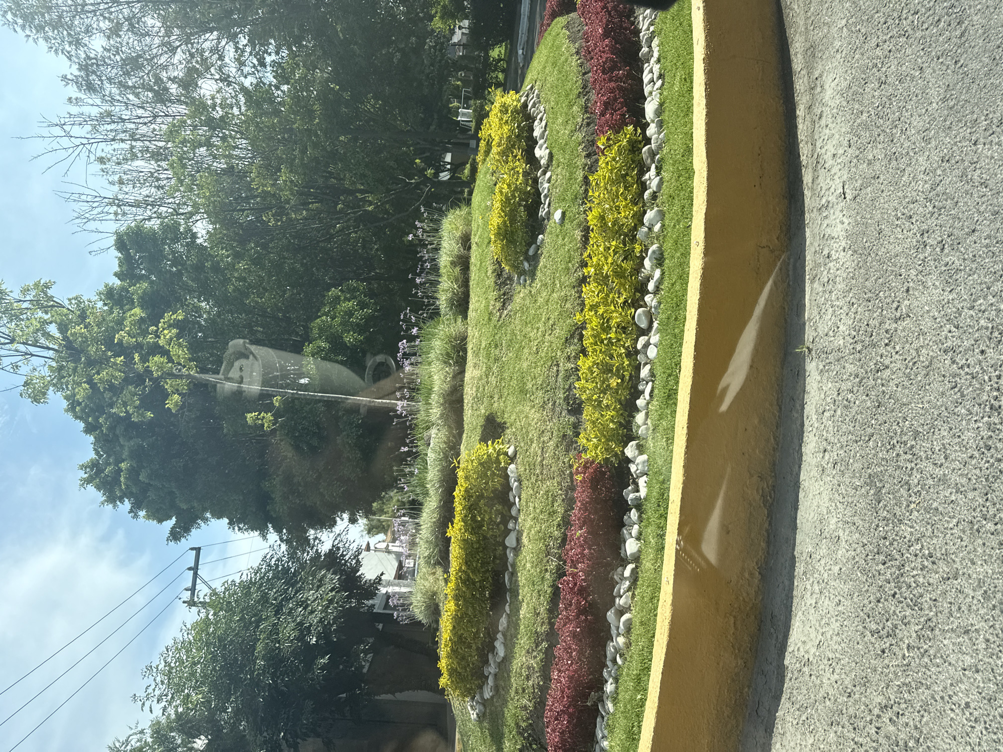 This media file presents an outdoor, daytime scene in San Bernardino Tlaxcalancingo, Mexico, primarily featuring a landscaped garden bed adjacent to a paved road. The garden is meticulously designed with several distinct rows of vegetation. These include stretches of bright green grass, vibrant yellow-leafed shrubs, and deep red-leafed shrubs, all separated by borders of smooth, white decorative stones. Further back in the garden, taller, grass-like plants with small purple flowers are visible. A large, ornate, dark-colored decorative pot or urn is partially visible among the greenery on the left side.
The garden bed is clearly delineated from the road by a prominent yellow concrete curb. The road surface is grey, coarse asphalt. In the background, numerous trees with dense green foliage fill the upper part of the frame, suggesting a lush environment. Above the treeline, a bright blue sky with some scattered clouds indicates a sunny day. Utility poles and power lines are discernible in the distance through the trees, hinting at a developed area. There are no people, explicit activities, or visible text in the image. The scene conveys a sense of peaceful, well-maintained public space under bright daylight conditions.