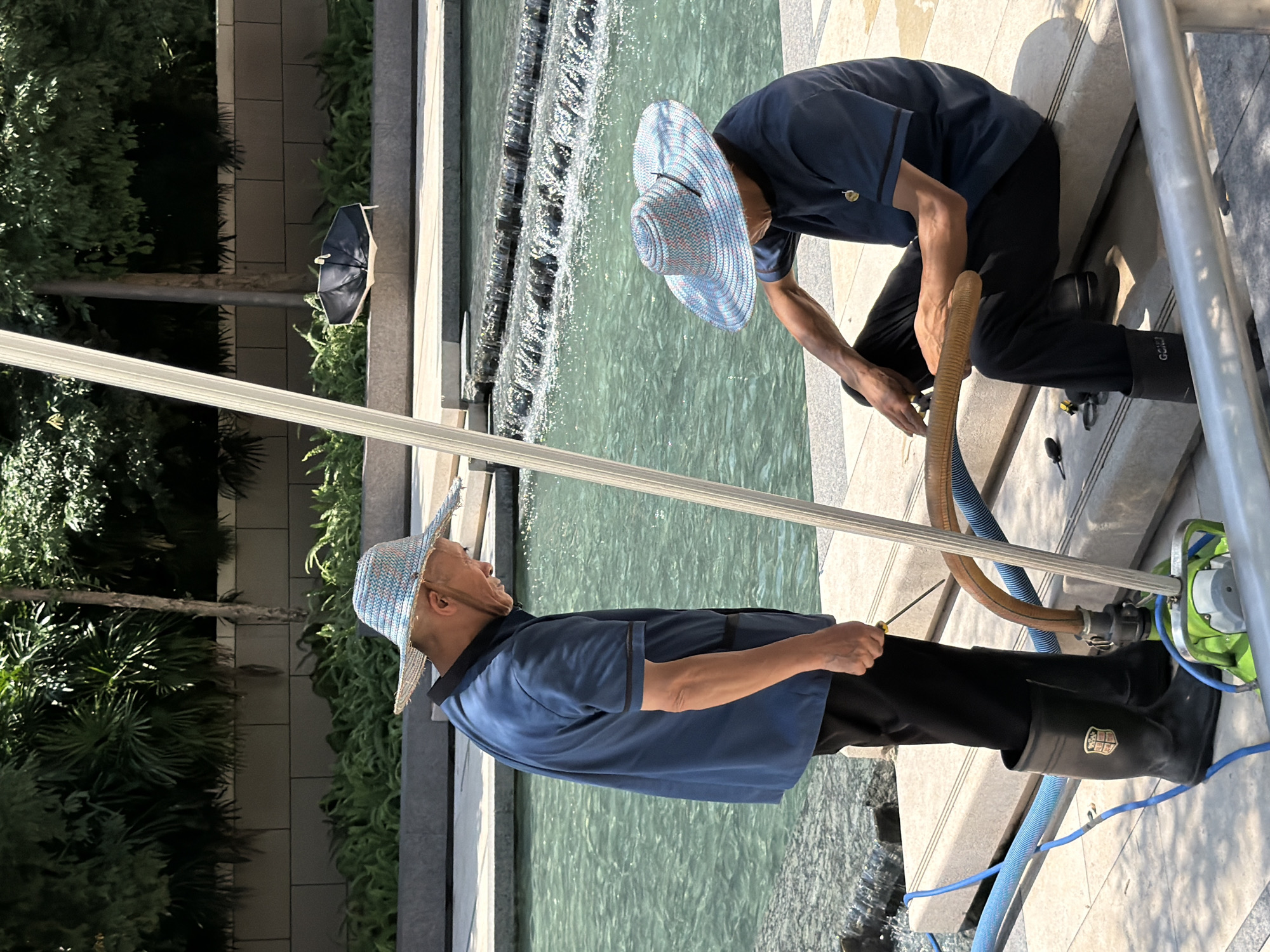 In Yuzhong District, China, two individuals are observed working near a body of water with cascading steps. The person on the left, wearing a blue short-sleeved shirt, black pants, and black rubber boots, is standing and looking down at the ground. They are holding a small tool in their right hand. The person on the right, also in a blue short-sleeved shirt and black pants, is seated on a stone ledge. They are wearing a wide-brimmed straw hat, as is the standing individual. Both are wearing similar straw hats with blue and pink checkered patterns. A flexible, corrugated hose and a blue hose are present, connected to a green piece of equipment that appears to be a pump. The water in the pool is a bright turquoise color, and the background features lush green foliage and a stone wall. A folded black umbrella rests on the stone wall in the background.