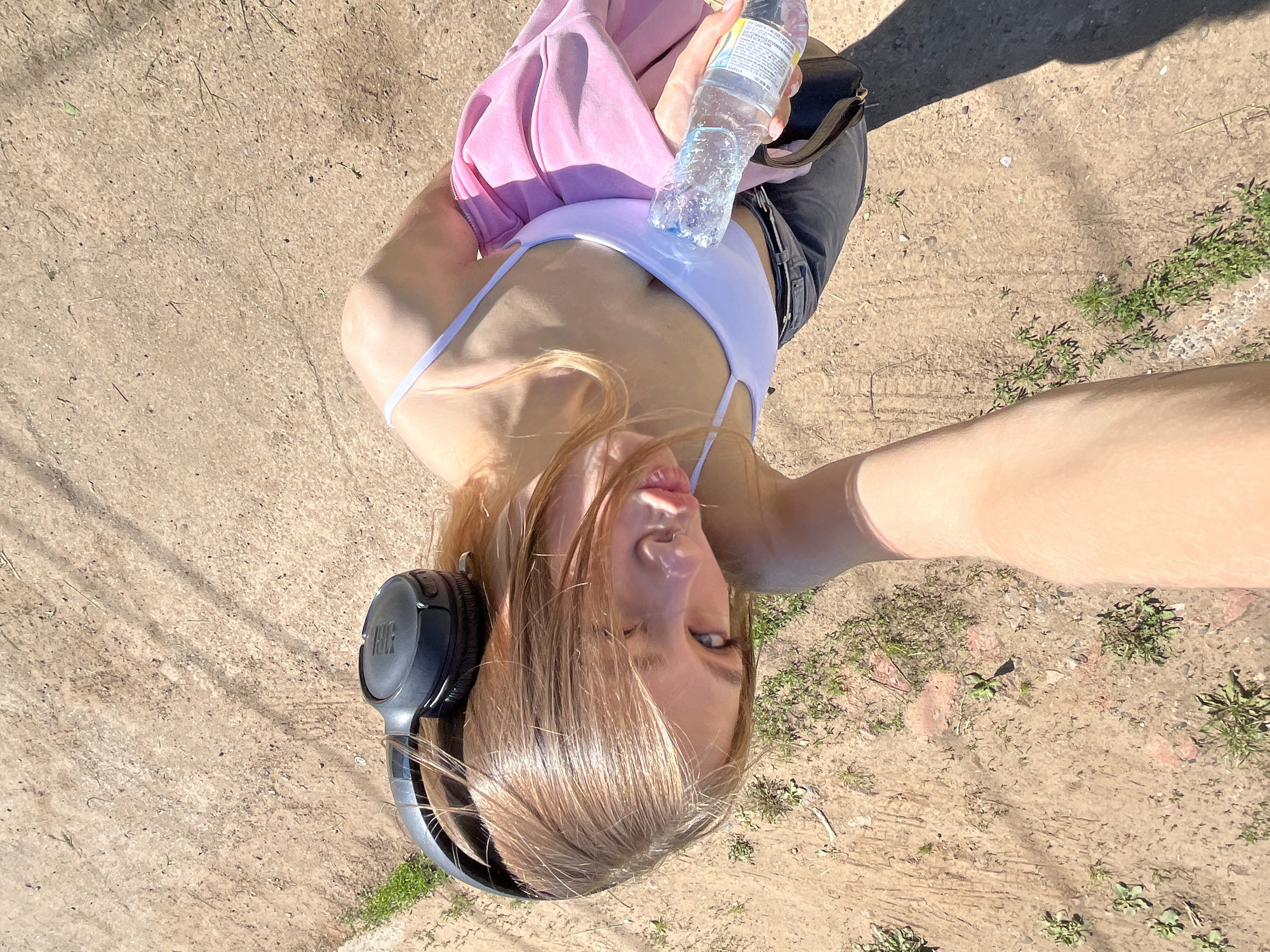 The image shows a person from a first-person perspective, holding a bottle of water. The person is wearing headphones and a pink jacket over a white top. The background appears to be a dirt or gravel path, with some sparse green vegetation. The lighting suggests it is daytime and sunny. The person's face is partially visible, looking towards the camera with a slightly pursed-lip expression. There is no specific indication of the location being in the city of Фоки, Russia.