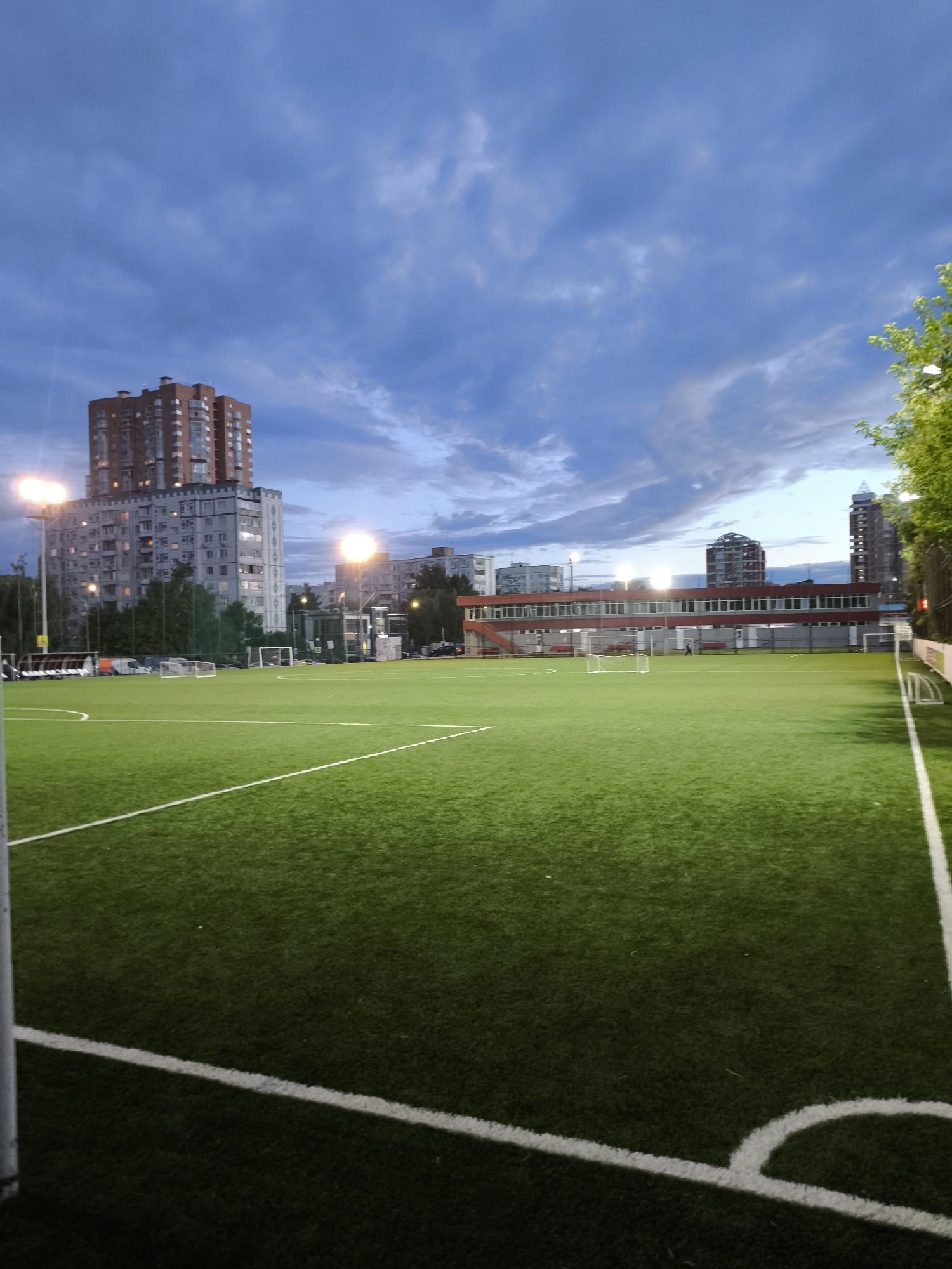 The image depicts an outdoor artificial turf soccer field at dusk. The sky is cloudy and a deep blue, suggesting the transition from day to night. Floodlights illuminate the field, casting a green glow on the turf. In the background, several multi-story buildings and a long, red-trimmed building with many windows are visible, hinting at an urban or suburban setting. A soccer goal is placed on the field, and white lines mark the boundaries and center circle of the playing area. There are no people actively engaged in a game, but the overall scene suggests a place used for sports and recreation. The lighting and cloudy sky create a dramatic and somewhat muted atmosphere.