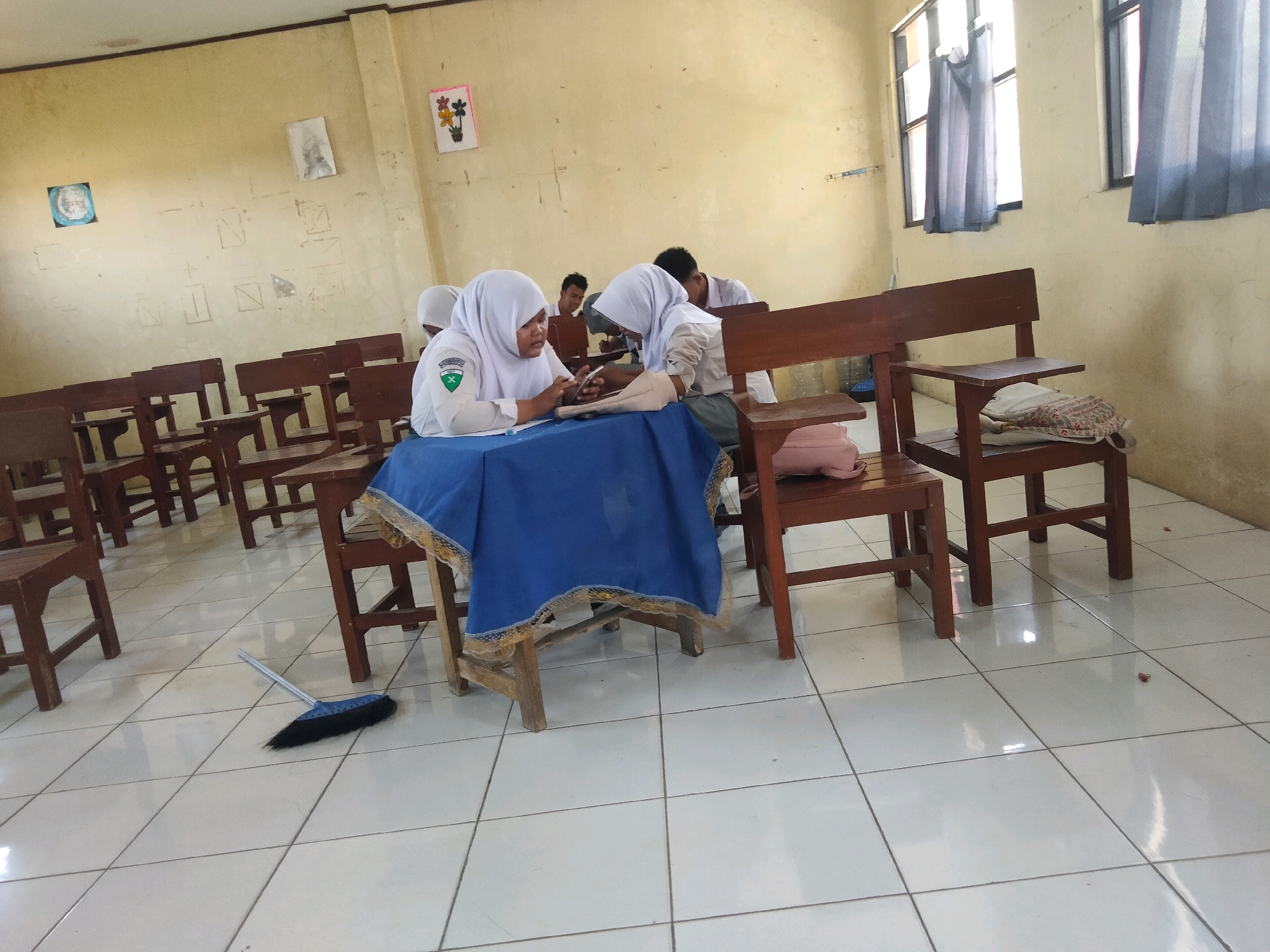 The image depicts an interior scene, likely a classroom, in Suka Sari, Indonesia. The room has a yellowish, textured wall and a white tiled floor. There are rows of wooden desks and chairs, some of which are occupied by students. In the foreground, two female students wearing white hijabs and school uniforms are seated at a desk covered with a blue cloth. They appear to be engaged in an activity, possibly studying or using a mobile phone. Another student, a male, is visible in the background, also seated at a desk. Various items, including a pink backpack and books, are scattered around the desks. A broom with a blue handle and black bristles is lying on the floor near the desks. Two windows with grey curtains are visible on the right side of the room. The overall impression is that of a casual learning environment within a school.