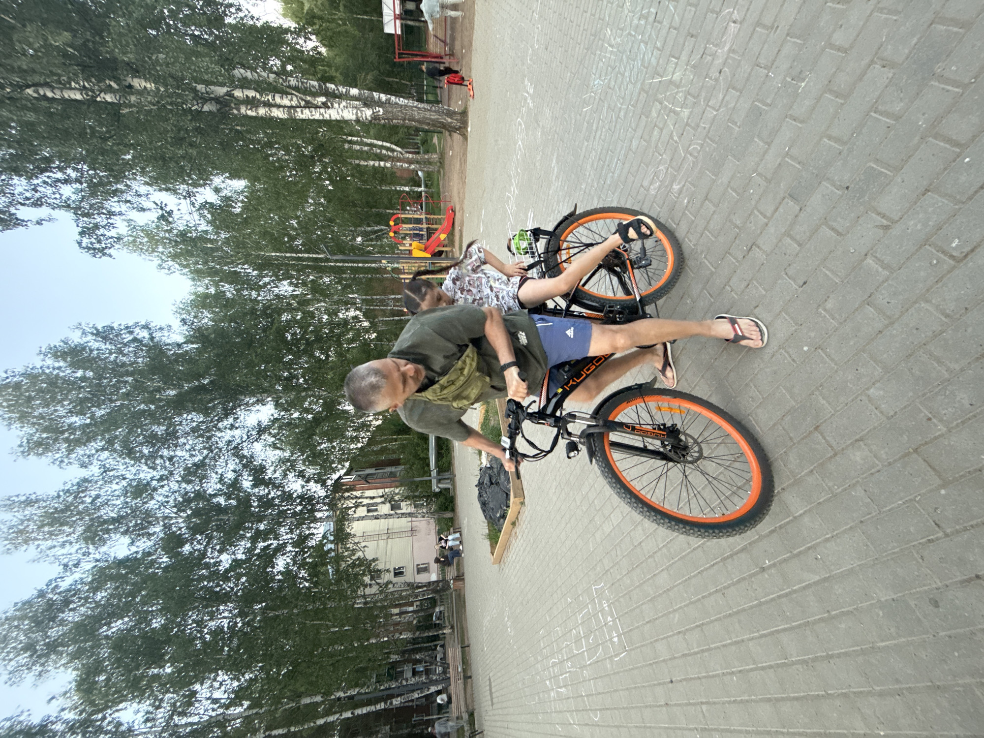 A man and a girl are interacting with a bicycle on a paved area in Votkinsk, Russia. The man, with gray hair and wearing a green shirt and blue shorts, is holding the bicycle's handlebars. The girl, wearing a patterned top and dark shorts, is sitting sideways on the bicycle's rear rack. The bicycle is black with orange rims. The paved area appears to be a walkway in a park, with birch trees and a playground visible in the background. There is a building in the distance, and the sky appears overcast.