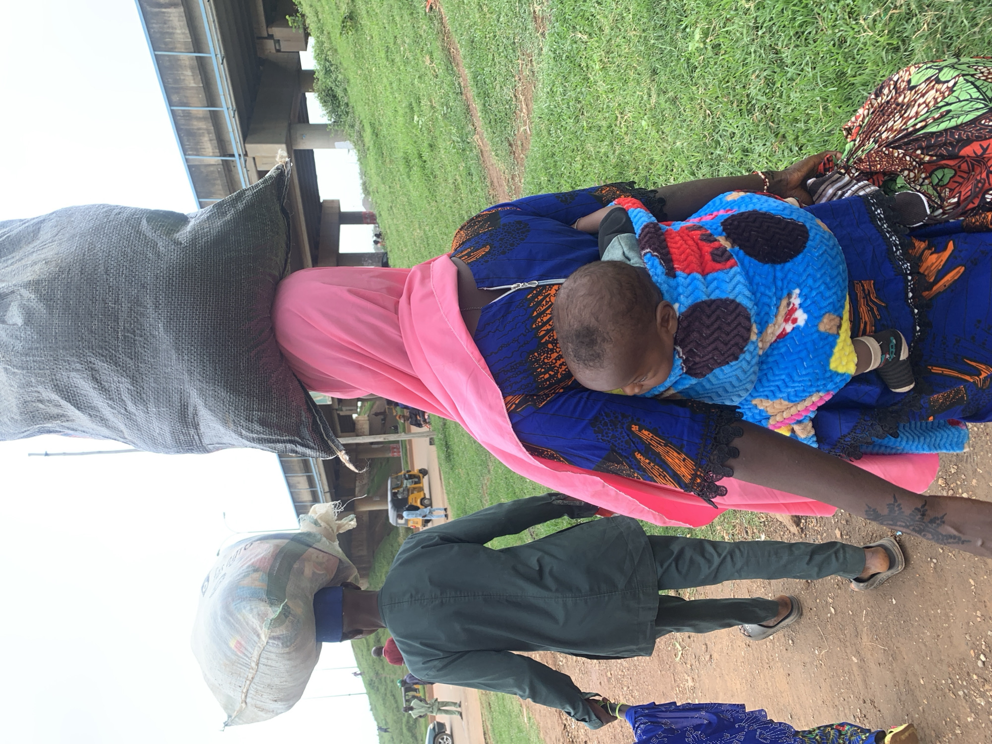 The image shows a group of people in what appears to be an outdoor market setting near Nyanya, Nigeria. In the foreground, a person is carrying a large sack of goods on their head. To their right, a woman is holding a baby wrapped in a colorful blanket. She is wearing a blue and orange patterned dress with black lace trim. Behind them, another person is seen from the back, wearing a dark green jacket and pants, also carrying a sack on their head.
The scene is set outdoors, with a grassy embankment to the left and a dirt path in the foreground. A concrete overpass or bridge is visible in the background, suggesting an urban or semi-urban environment. The sky is overcast, indicating it might be a cloudy day. The presence of a yellow motorized rickshaw in the background further suggests a location in Nigeria.
The overall impression is one of daily life and commerce, with people engaged in the activity of transporting goods and caring for children. There is no visible text in the image. The time of day is not explicitly discernible, but the lighting suggests it is daytime. The mood is neutral, depicting ordinary activities without any strong emotional cues.
