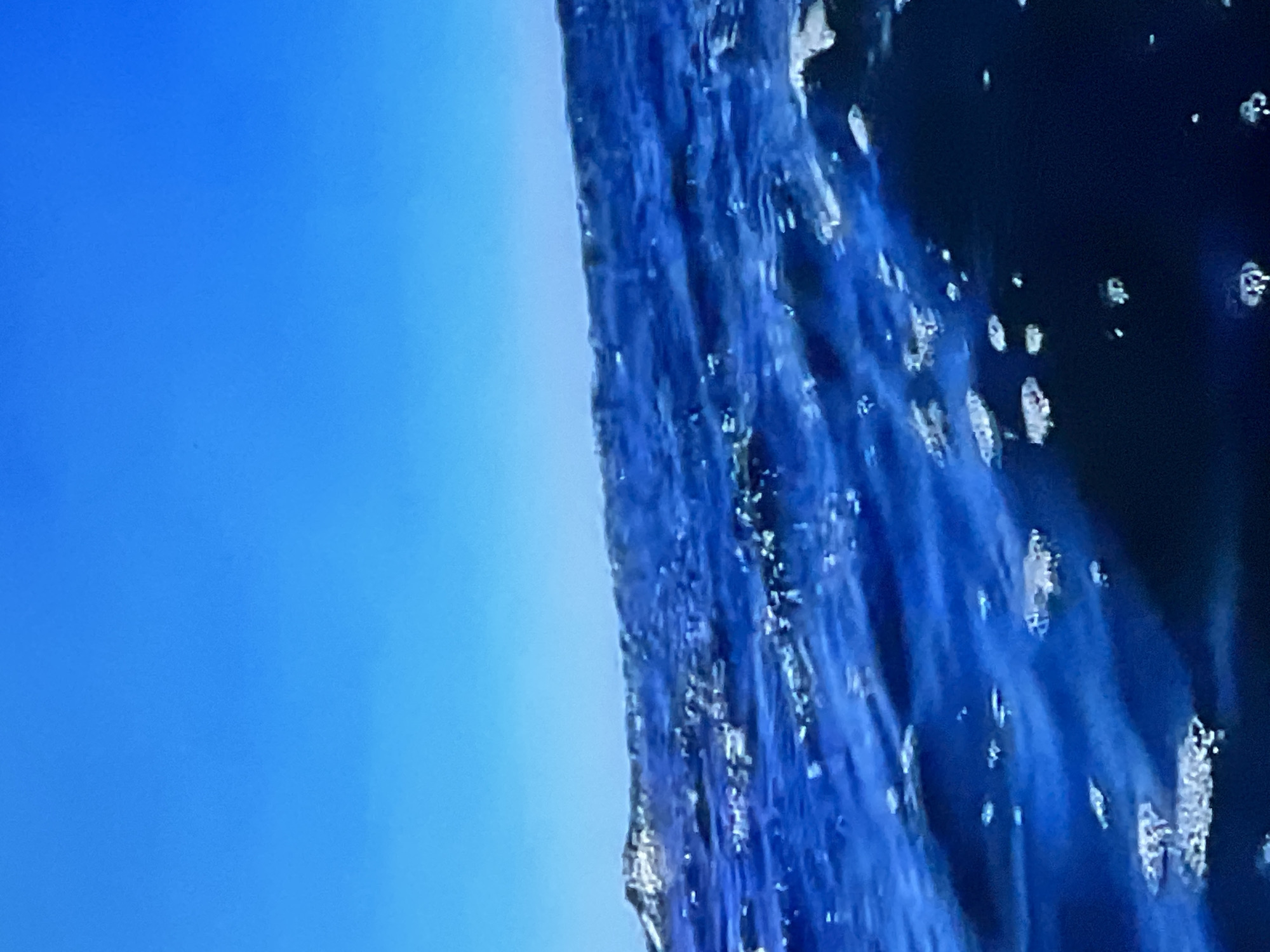 The image shows a close-up view of the ocean with a clear blue sky. The water is a deep blue with white foam on the surface, indicating movement and possibly waves. The sky is a solid, bright blue with no clouds visible. There is no indication of the city of Casablanca, Morocco, or any people or objects within the scene.