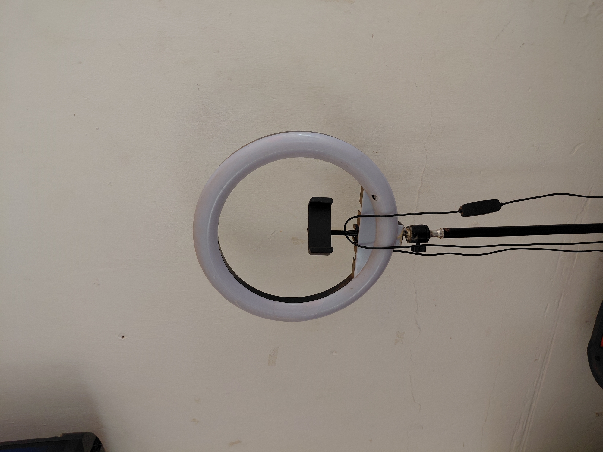 The image displays a close-up, slightly tilted view of a ring light set up against a light-colored, textured wall. The ring light, which appears to be off, is predominantly white and circular, with a black phone holder mounted centrally within its aperture. Several black cables extend from the right side of the ring light. One cable connects to a black, elongated inline controller, likely for power or brightness adjustment, before continuing off-frame to the right. The ring light is attached to a black pole or arm, suggesting it's mounted on a stand or tripod, only a portion of which is visible.
The wall behind the ring light is a neutral, possibly off-white or very light beige color, showing visible texture, minor imperfections, faint cracks, and some smudges or discoloration across its surface. This suggests an indoor setting, possibly a room in a home or studio. In the bottom left corner, a dark, angular object, possibly part of an electronic device like a speaker or computer, is partially visible. Another dark object with an orange or red accent is partially visible in the bottom right corner, indicating additional equipment nearby. The overall lighting is soft and ambient, and no specific time of day or weather can be discerned. There is no visible text in the image. The scene suggests a setup for vlogging, video conferencing, or photography.