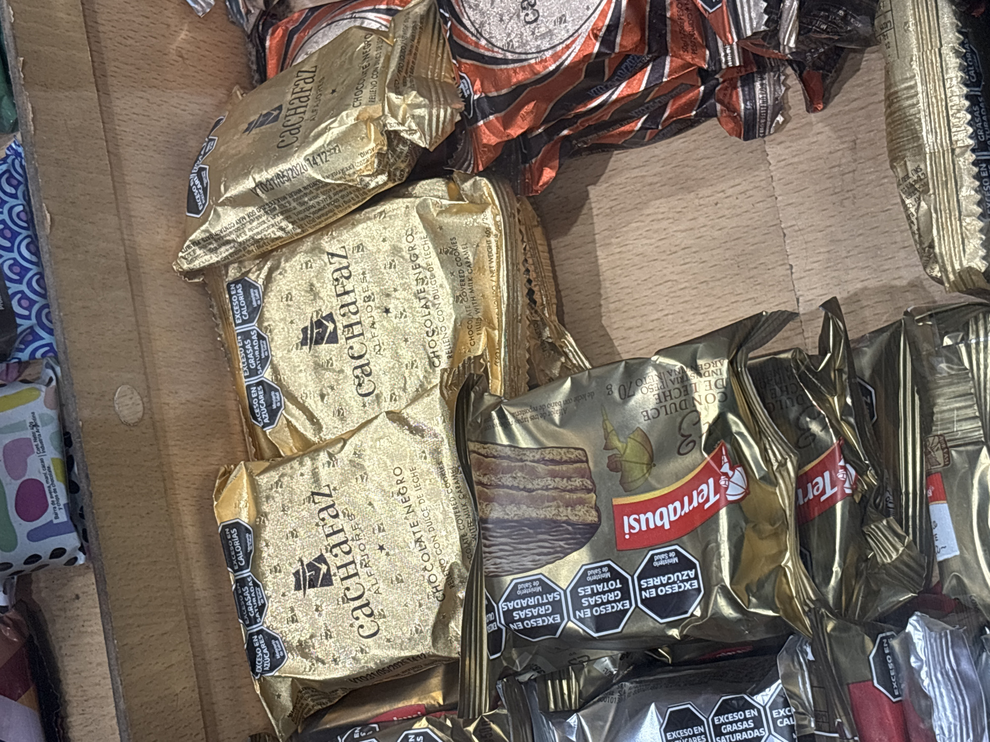The image shows a close-up view of a pile of individually wrapped alfajores, a traditional Argentinian confection. Several brands are visible, including "Cachafaz" and "Terrabusi." The alfajores are packaged in gold and black wrappers, with some wrappers featuring warning labels such as "Exceso en grasas saturadas" (Excess in saturated fats), "Exceso en azúcares" (Excess in sugars), and "Exceso en calorías" (Excess in calories). The products are placed on a wooden surface. Some alfajores are partially obscured by others, creating an overlapping arrangement. No people are present in the frame. The overall impression is of a collection of packaged snacks, likely photographed indoors.