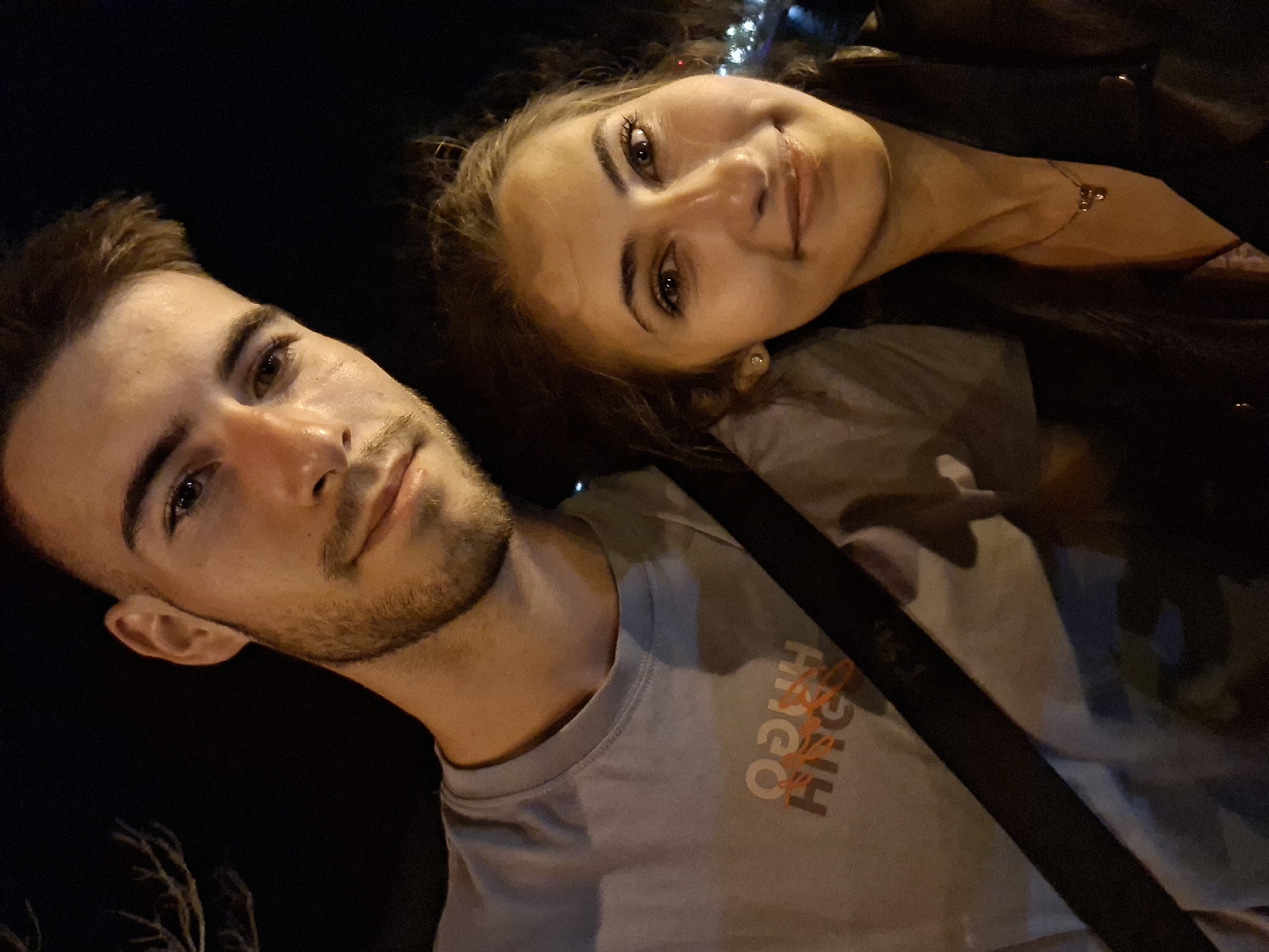 A nighttime, close-up selfie shows a young man and a young woman. The man is on the left, his face turned towards the camera, and the woman is on the right, also facing the camera. Both are smiling. The man is wearing a light gray t-shirt with an orange graphic print. The woman is wearing a dark jacket over a lighter-colored garment and a necklace. The background is dark, with some blurred lights visible in the distance, suggesting an urban setting.