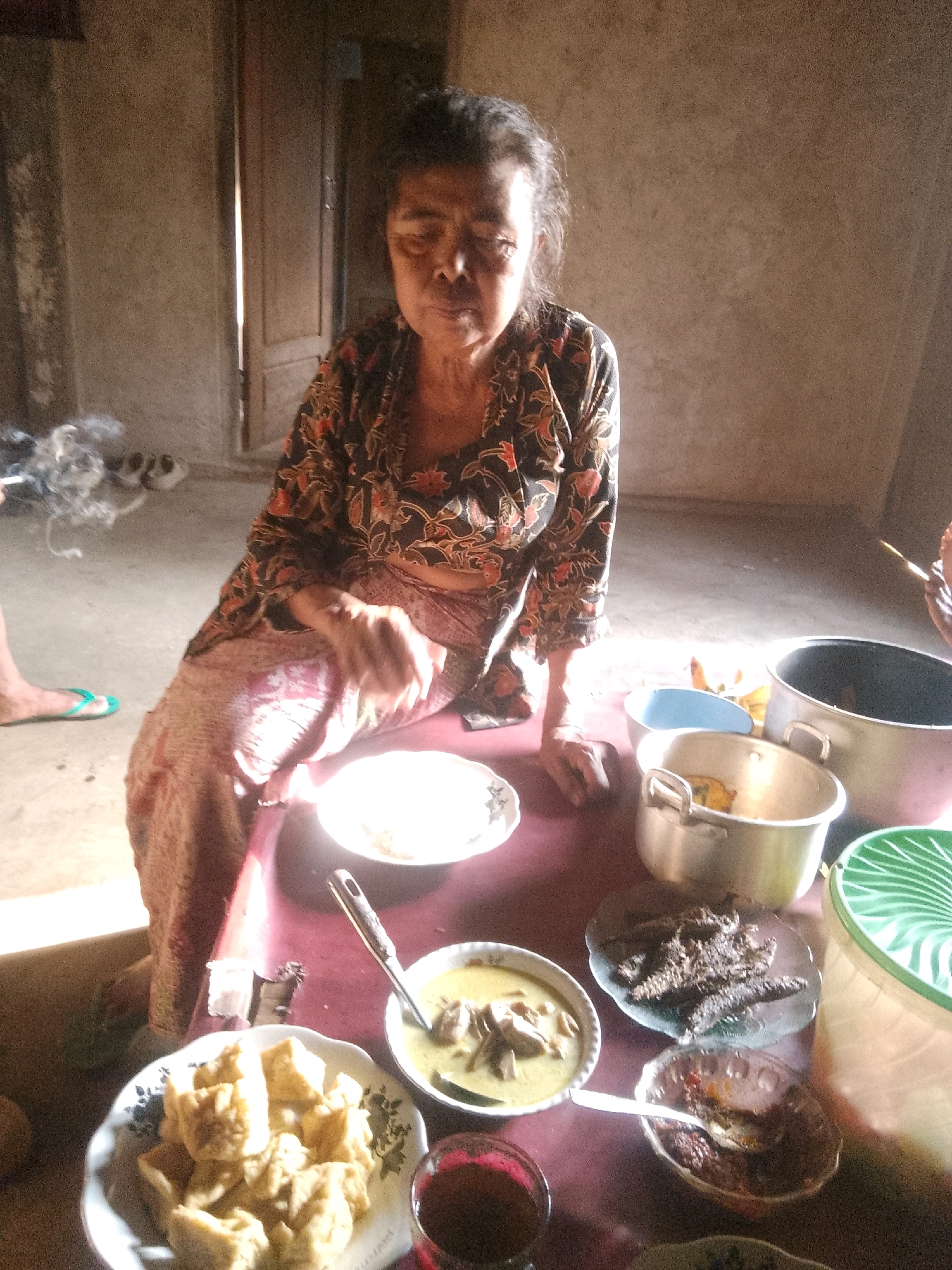 A middle-aged Indonesian woman sits at a table in a dimly lit room. She wears a traditional batik-patterned shirt and skirt. In front of her is a plate of food. The table is laden with several bowls and pots of food, suggesting a meal is being served. The room appears to be a simple dwelling, with bare walls and a wooden door in the background. Smoke from a cigarette drifts in the air near the woman. The overall scene depicts a candid moment of everyday life in Krucil, Indonesia.