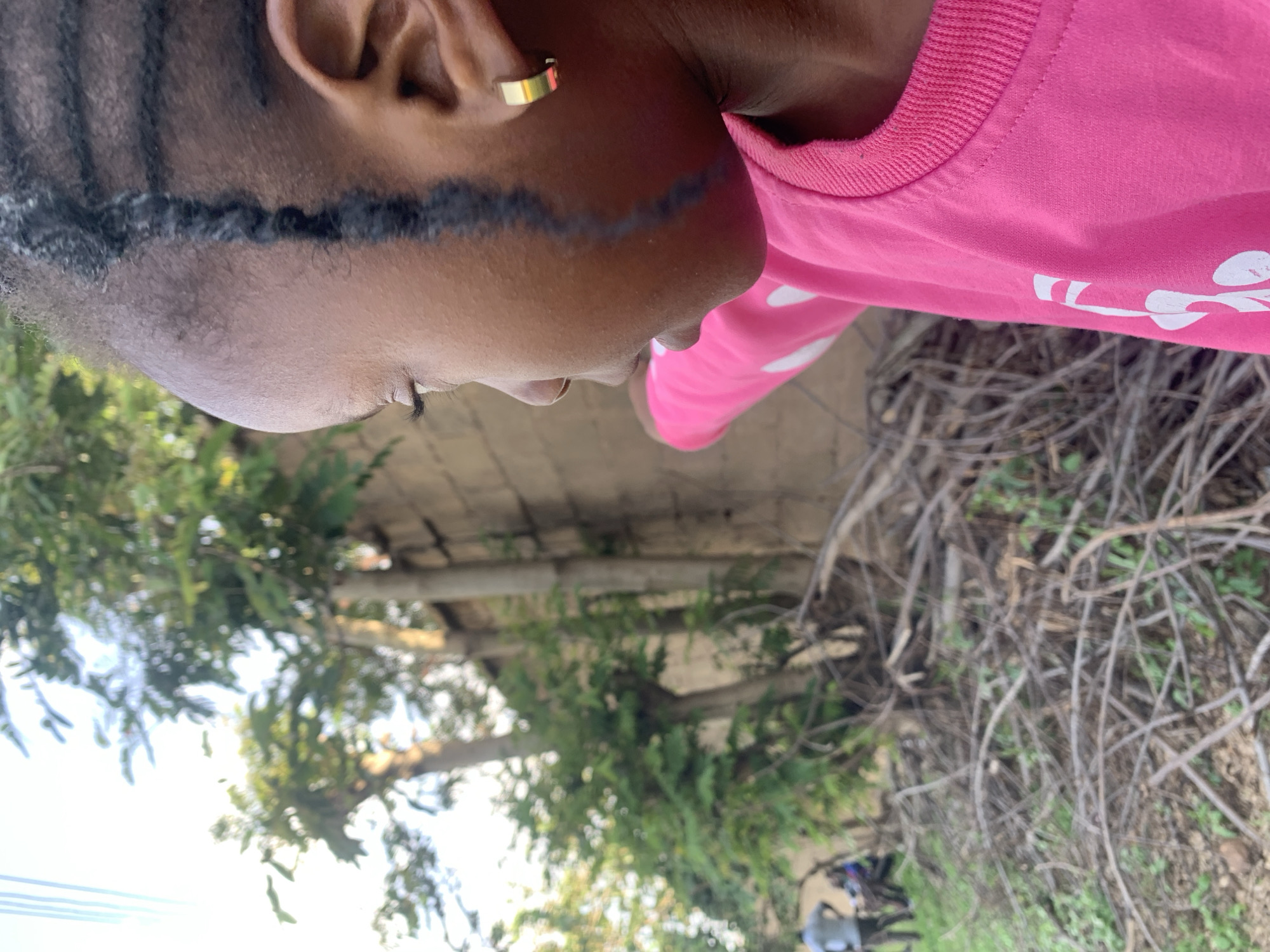 In Jos, Nigeria, a close-up, profile shot captures a young Black girl wearing a bright pink sweatshirt adorned with white polka dots and other white designs. She has braided hair and a gold hoop earring in her right ear. Her gaze is directed to the left, with her right arm outstretched in that direction. The background reveals a rustic outdoor scene. To the left of the girl, lush green trees and vegetation are visible. Behind her, a tan-colored wall of a building stands, with more greenery peeking from behind it. The ground in the foreground is covered with a dense layer of dry, brown branches and twigs, interspersed with patches of green grass. In the far left of the image, two individuals, seemingly adults, are visible in the background, one standing and the other appearing to be seated. The overall impression is an outdoor environment, possibly a yard or undeveloped area within the city of Jos.