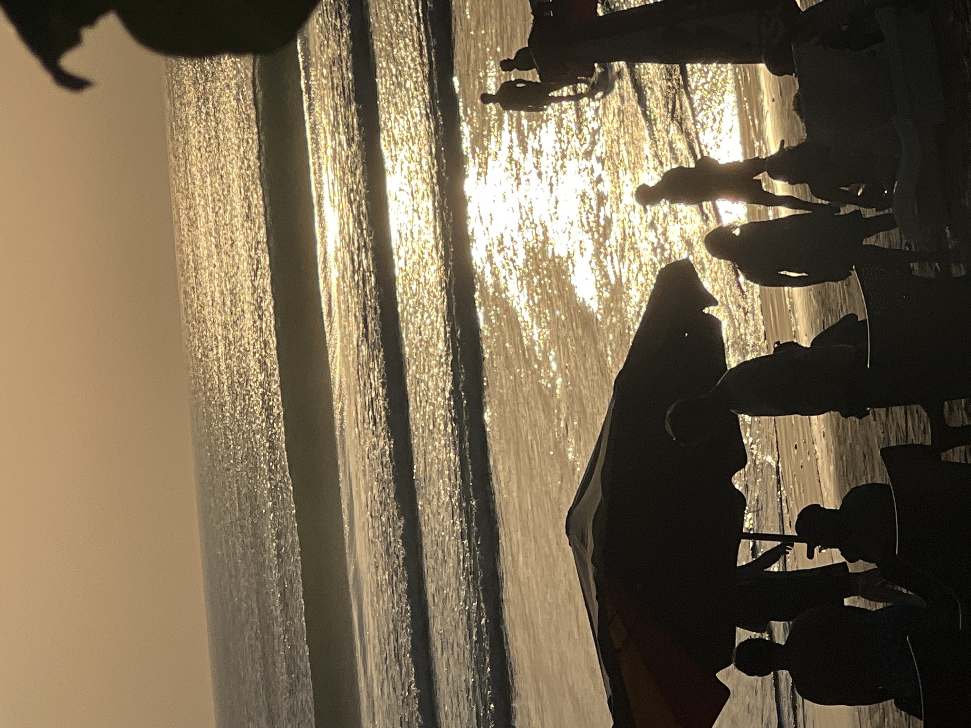 At sunset on Kuta Beach in Bali, Indonesia, several people are visible on the sand and in the shallow ocean. The sun's reflection creates a golden shimmering path across the water, with waves gently rolling towards the shore. In the foreground, silhouettes of people are gathered under a large beach umbrella. Some are seated in chairs, while others stand. Further out in the water, more figures are wading or standing, their forms outlined against the bright reflection of the setting sun. The overall scene conveys a sense of peaceful evening relaxation by the sea.