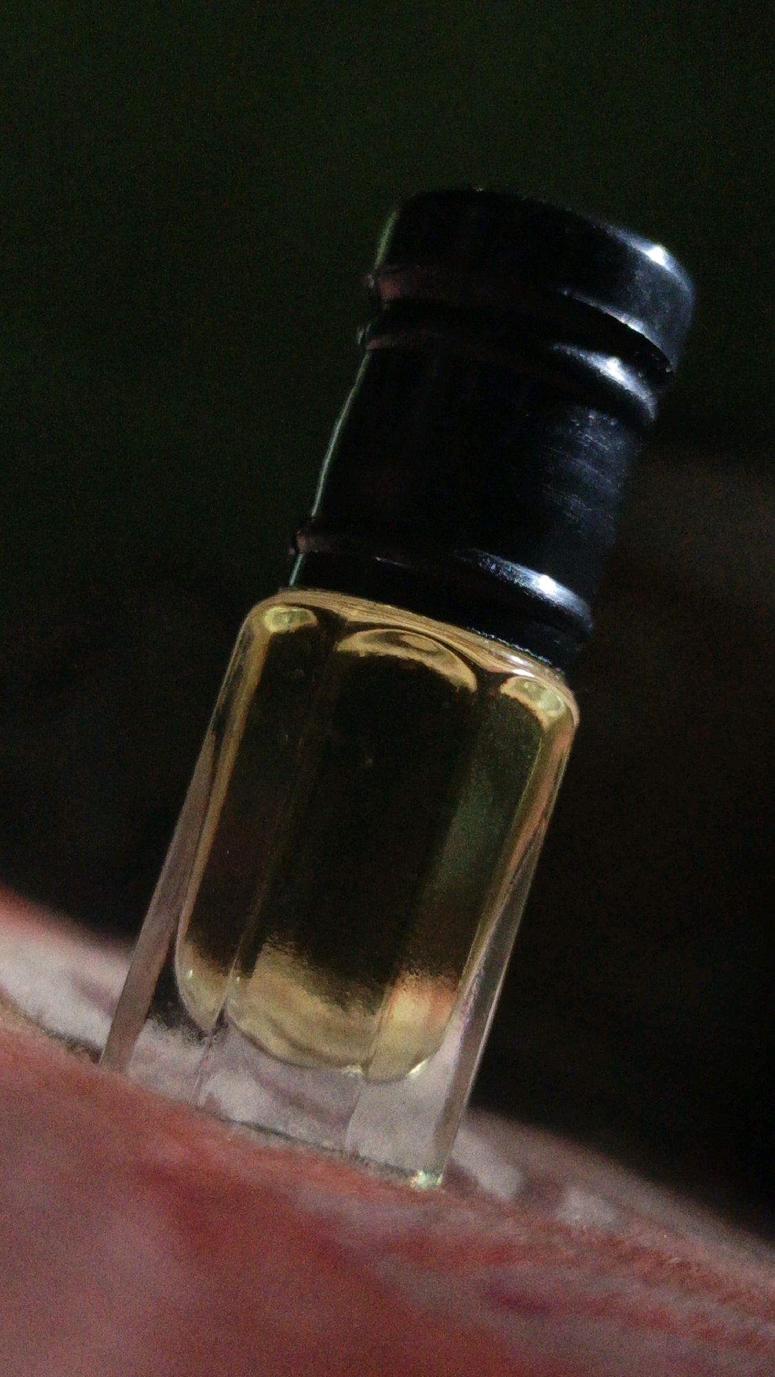 A close-up, low-angle shot shows a small, clear glass bottle with a black cap filled with a golden liquid, possibly perfume or oil. The bottle is tilted and rests on a reddish-pink surface, which appears to be skin. The background is dark and out of focus, with a hint of green. The lighting is dramatic, creating highlights and shadows on the bottle and liquid. The image is shot in Nabinagar, Bangladesh.
