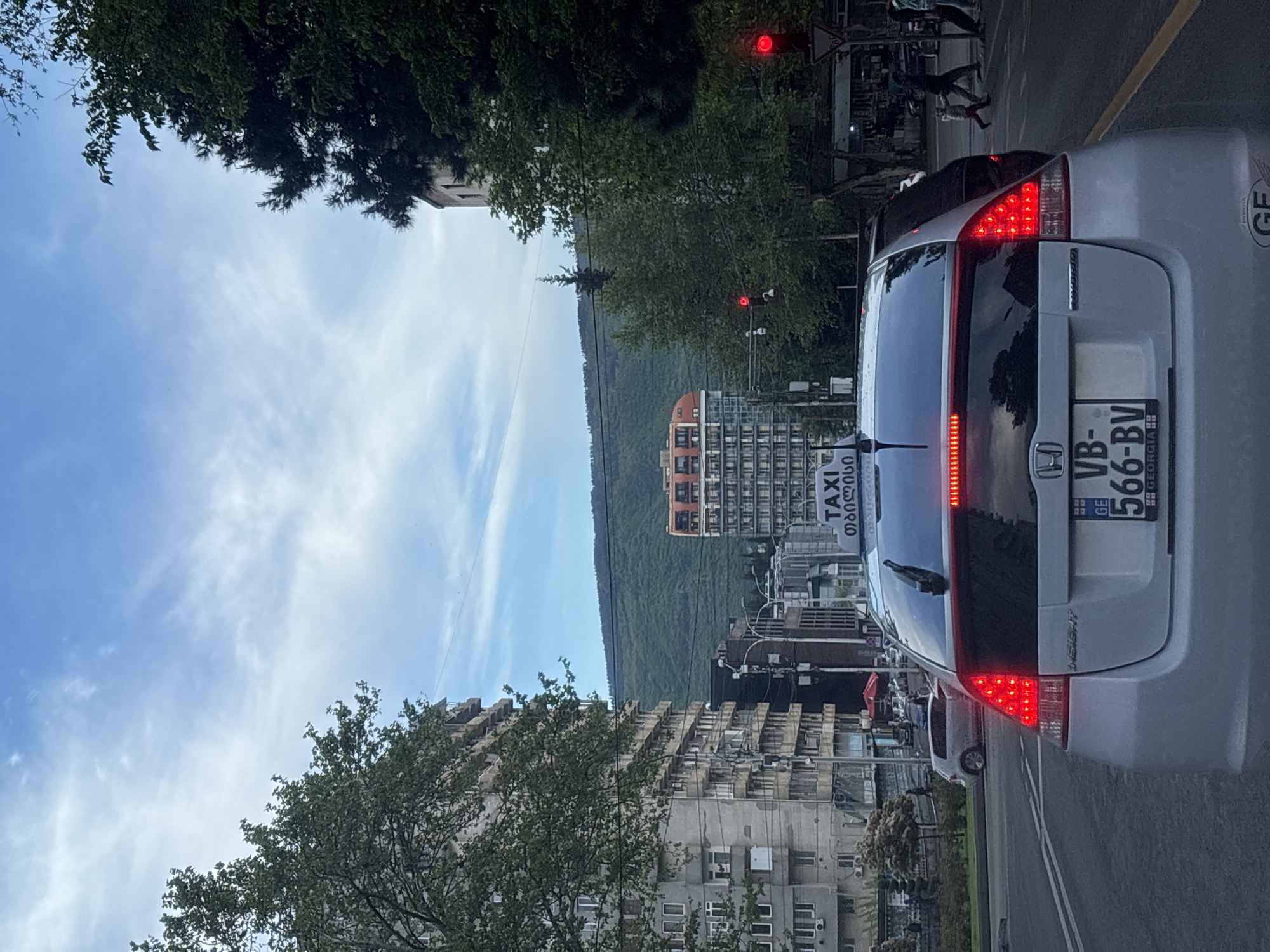 The image captures a street scene in Tbilisi, Georgia. A silver car with the license plate "VB-566-BV" is in the foreground, displaying a "TAXI" sign in Georgian script. The car is stopped at a traffic light, which is red. Behind the car, the street stretches into the distance, flanked by buildings of varying heights and architectural styles, including older concrete apartment blocks. A tree with green foliage is visible on the left side of the frame. In the background, a green mountain range is visible under a partially cloudy blue sky. Pedestrians can be seen crossing the street in the distance.