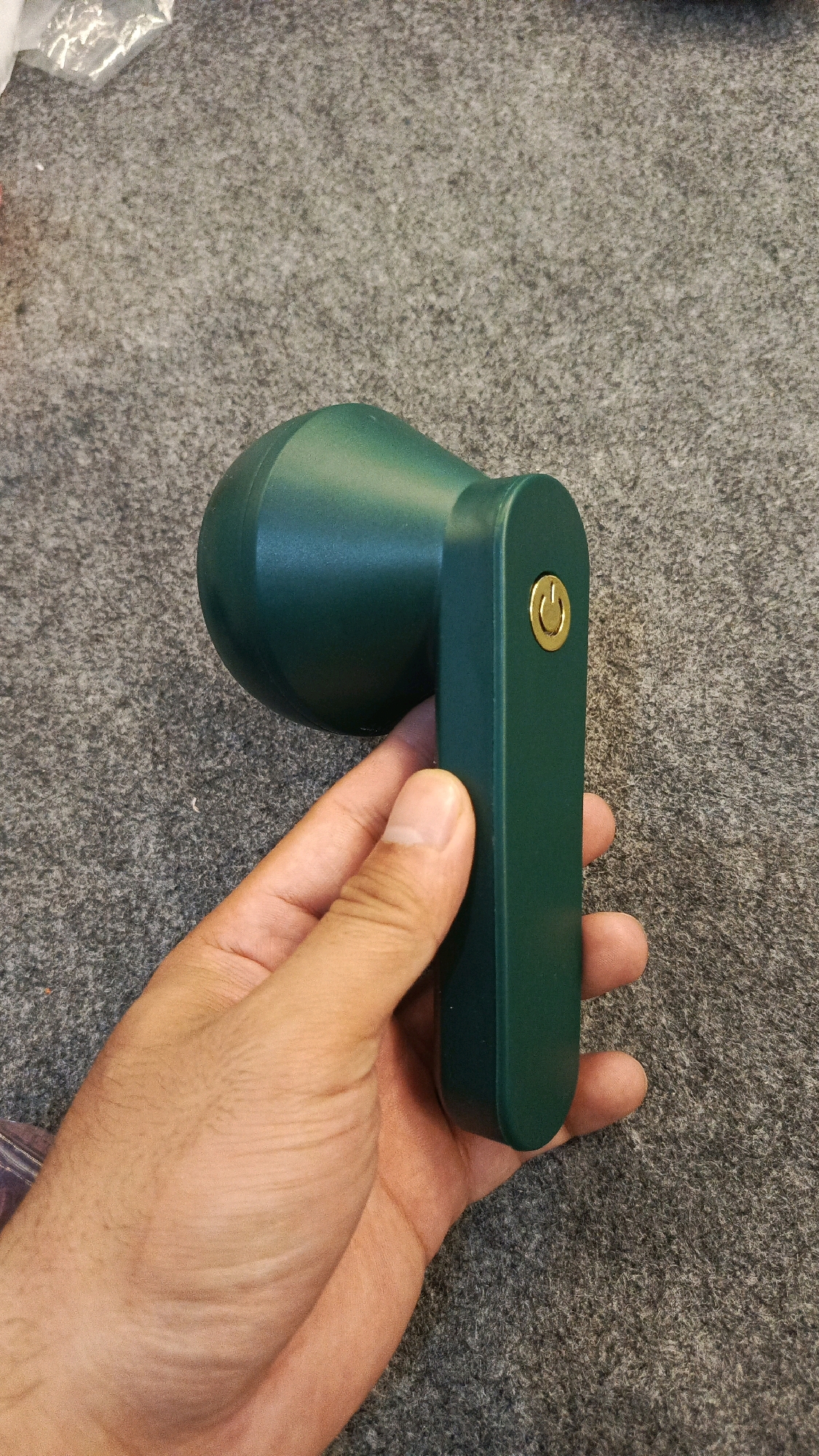 A person's hand is holding a dark green fabric shaver with a gold power button against a grey carpeted surface. The fabric shaver has a rounded head and a rectangular handle. The background is out of focus and shows a grey, textured surface. There are no discernible landmarks or contextual clues that would indicate a specific location in Babadan, Indonesia.