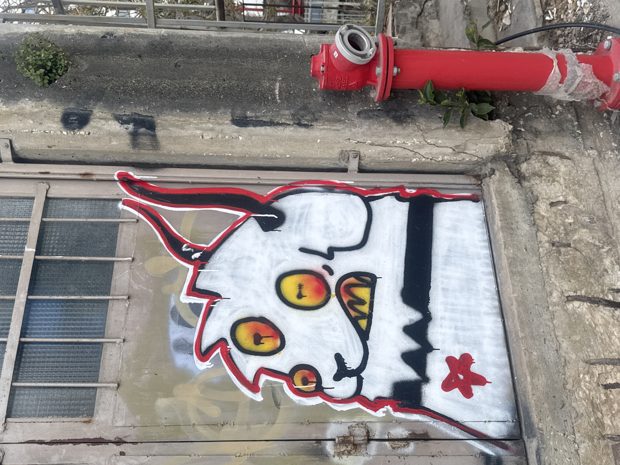 A colorful graffiti mural of a whimsical monster-like character is painted on a metal door or hatch, positioned below a gray concrete ledge. A red fire hydrant is affixed to the ledge above the mural. The mural features a white-faced cartoon character with red outlines, large yellow eyes, a black nose, and a sharp-toothed grin. A wide black stripe runs vertically through the character's body, and a red star-shaped mark is painted near the bottom right corner of the mural. The door is divided into several panes by metal bars, and there are hints of other graffiti beneath the main mural. The surrounding environment appears to be an urban setting. This photo was taken in Tel Aviv, Israel.