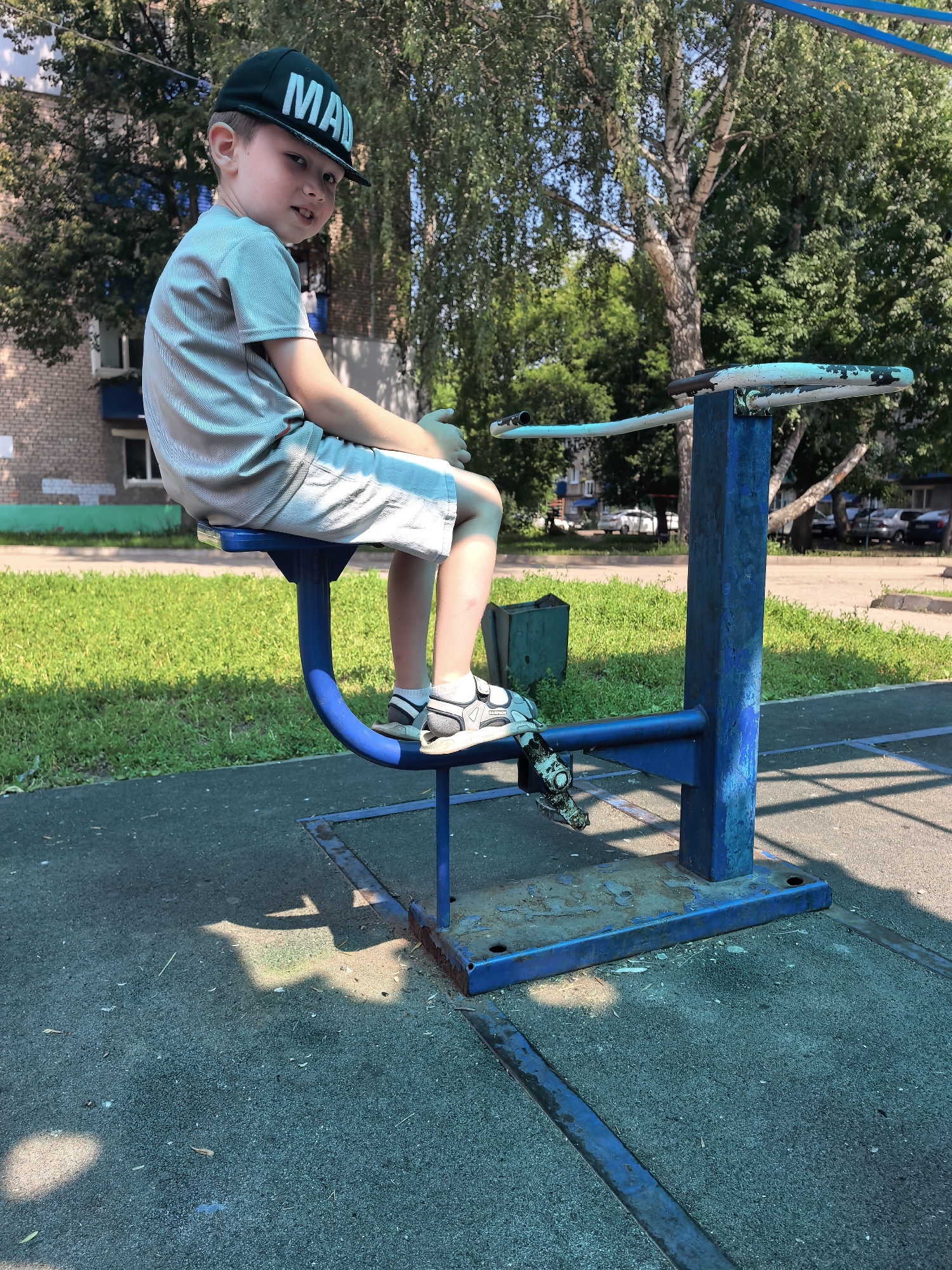 The image shows a young Caucasian boy, appearing to be around 6-8 years old, sitting on a blue outdoor exercise machine. He is wearing a grey t-shirt, grey shorts, a dark green baseball cap with white lettering that reads "MAD", and black and white sandals. The boy is looking towards the camera with a slight smile.
The exercise machine is blue and made of metal, with a seat and footrests. It is situated on a dark grey rubberized playground surface. In the background, there is a grassy area with trees and a brick building with blue trim. Cars are visible parked in the distance. The scene appears to be a public playground or park.
Based on the architectural style of the building and the type of playground equipment, this scene is consistent with urban public spaces found in cities like Sterlitamak, Russia, particularly in residential areas. The presence of trees, a multi-story apartment building, and common playground equipment suggests a typical urban park or schoolyard setting. The overall lighting indicates it is daytime, likely late morning or early afternoon, given the strong shadows cast on the ground.