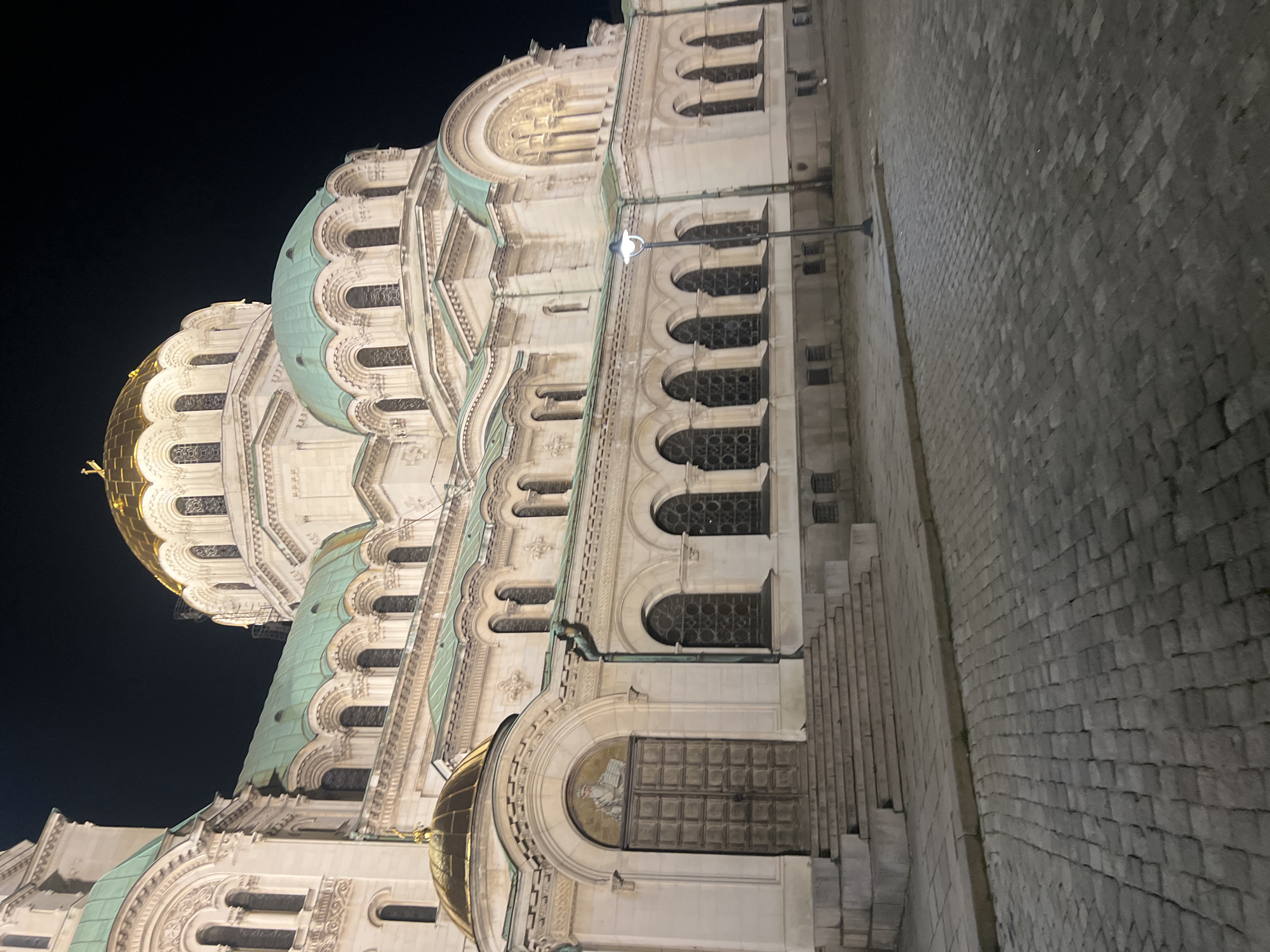 The image shows the exterior of the Alexander Nevsky Cathedral in Sofia, Bulgaria, at night. The cathedral is a large, white Orthodox cathedral with multiple domes, one of which is gold and illuminated. The facade features numerous arched windows and decorative architectural elements. In the foreground, a cobblestone street and a wide set of stone stairs leading to a large wooden door are visible. A streetlamp stands on the right side of the image. The sky is dark, indicating nighttime. There are no people or other subjects visible in the frame. The scene is static and captures the grandeur of the cathedral illuminated against the night sky.