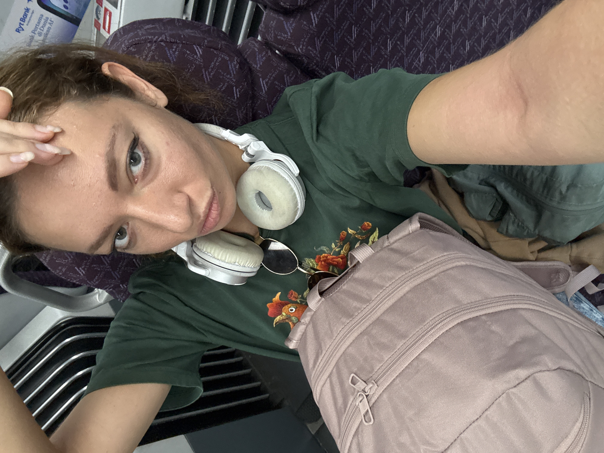 A woman with light skin and light brown hair is seated on a train, taking a selfie. She is wearing a dark green t-shirt with an embroidered rooster and floral design, and a pair of white headphones around her neck. A light pink backpack rests on her lap. She has dark eyeliner on her eyes and is puckering her lips. The background shows the interior of a train car with purple patterned seats and metal luggage racks. The image appears to be taken in Sepang, Malaysia, as indicated by a sign visible in the background that says "Ryt Bank."