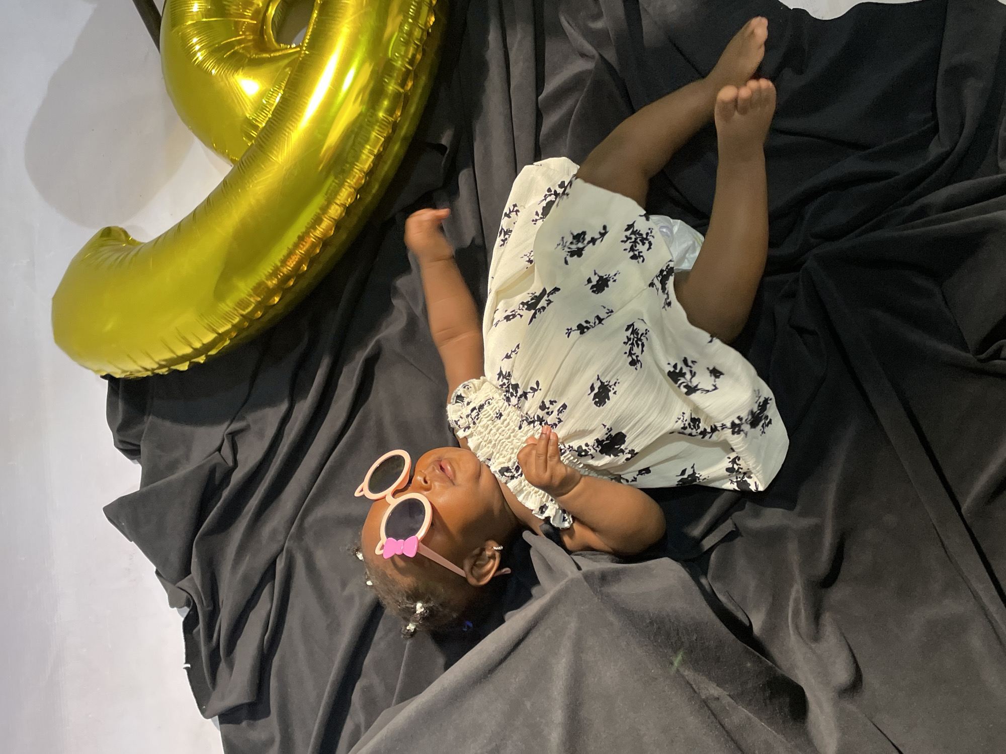 A baby lies on their back on a dark grey cloth. The baby is wearing a light yellow dress with black floral patterns and pink sunglasses. A gold balloon number six is to the left of the baby. The background is a light grey wall. The location is Jos, Nigeria.