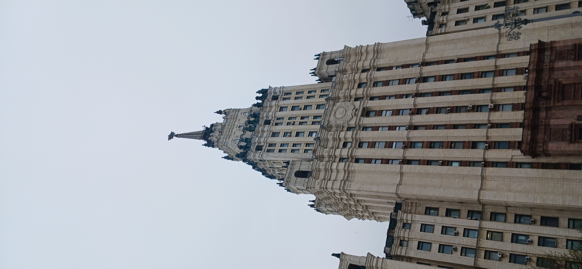 The image shows a large, ornate building in an unknown city, viewed from below. The building has many windows and architectural details, including a spire at the top. The sky is overcast. The building appears to be located in Russia.