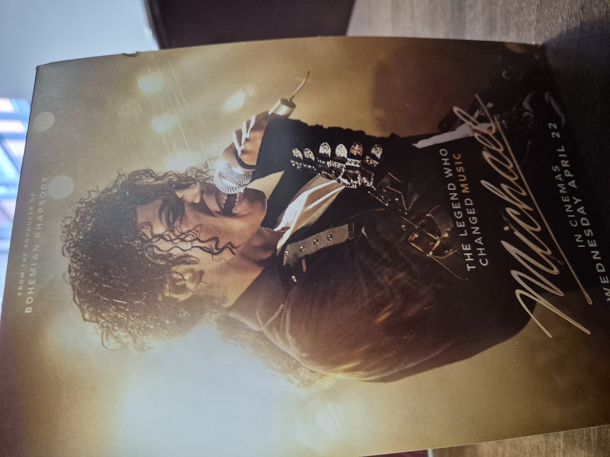 The image shows a movie poster for "Michael," a film about Michael Jackson, "the legend who changed music." The poster has a picture of Michael Jackson singing into a microphone. Text on the poster also mentions "From the Producer of Bohemian Rhapsody" and "In Cinemas Wednesday April 22." The poster appears to be lying on a wooden surface, possibly a table. The location in West Suffolk, United Kingdom is unknown.