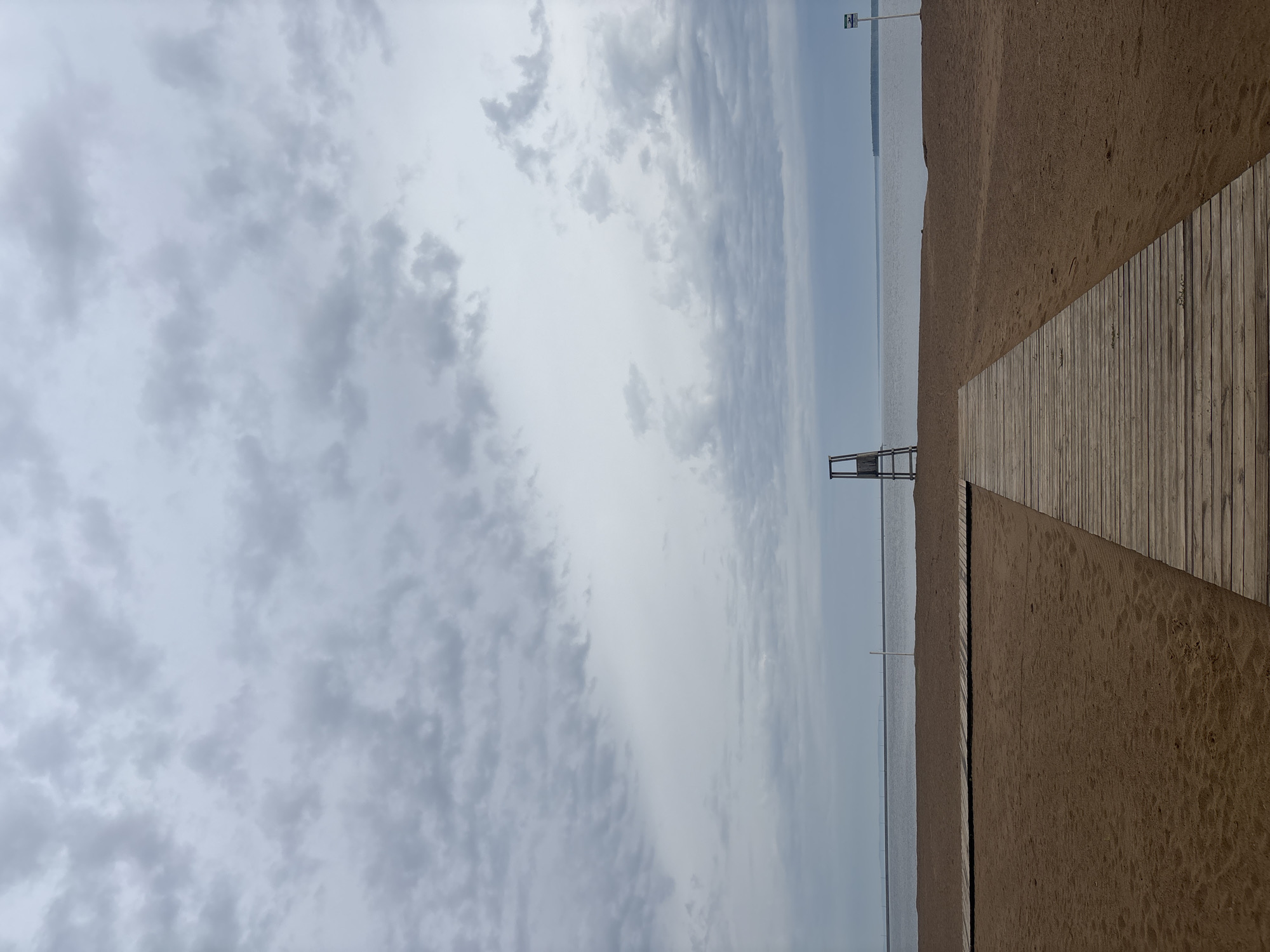 The image shows a beach scene in Chaikovsky, Russia. A wooden boardwalk extends from the right edge towards the horizon. To the left of the boardwalk is a sandy beach leading to calm water. In the distance, a lifeguard stand is visible. The sky is overcast with dense, layered clouds. There are no people visible.