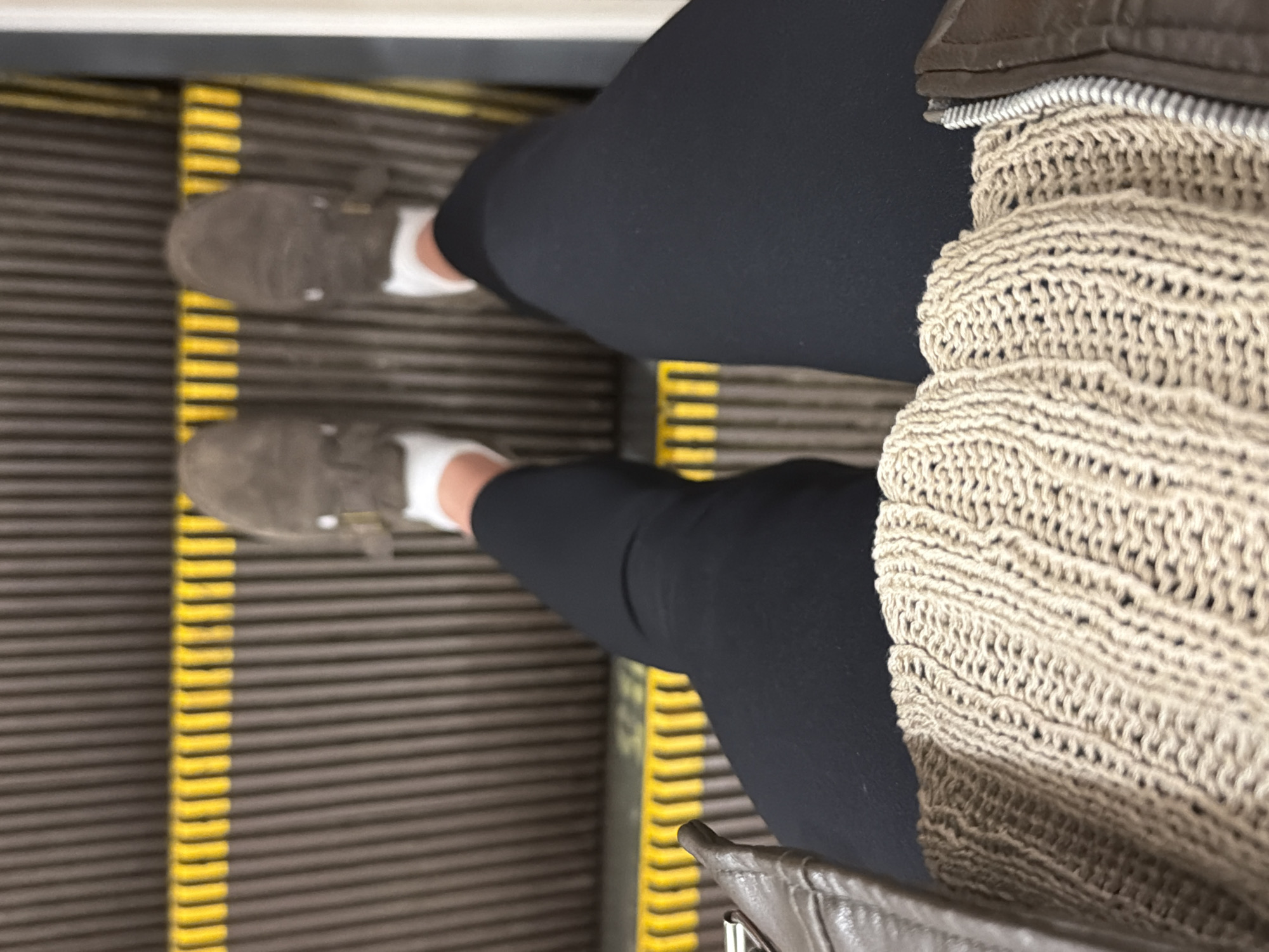 The image, captured from a high-angle, first-person perspective, shows a person riding an escalator. The subject's legs are prominent, clad in dark black leggings or pants, paired with white socks and gray-brown suede-like flat shoes or moccasins. Their feet are positioned on the textured, dark gray escalator steps, which are delineated by bright yellow horizontal safety lines along their edges.
Part of a light beige or tan knitted garment, possibly a sweater or cardigan, with a distinct open-weave pattern, is visible on the right side of the frame, along with a brown, possibly leather, detail that could be part of a jacket or bag.
The setting is an indoor escalator, suggesting a public space like a metro station, shopping mall, or airport. Given the provided context, this location is in Moscow, Russia. The lighting appears artificial and uniform, typical of an indoor environment. No specific time of day or external weather conditions can be discerned. There is no visible text within the image. The scene depicts a routine moment of transit or travel.