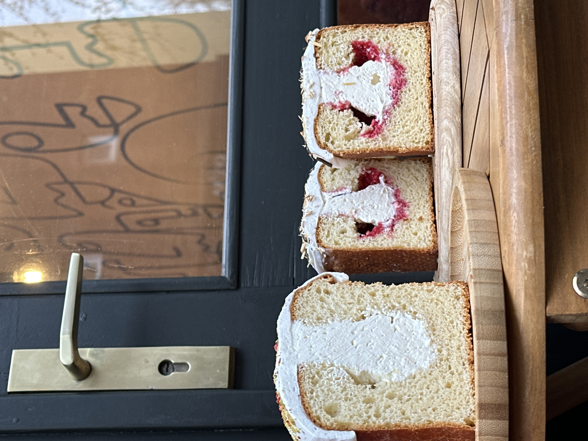 The image shows three slices of cake, stacked vertically and held in a wooden holder. The cake appears to be a brioche type, with a light-colored crumb and a slightly browned crust. Each slice is filled with a white cream, and the top two slices also have a red, jam-like filling.
The cake slices are positioned in front of a dark blue door or frame with a brass handle and keyhole. Reflected in the glass of the door is a drawing or design.
The location appears to be the "MOM café" in Novi Sad, Serbia, based on the appearance of the wooden holder and the style of cake which aligns with photos and descriptions of offerings from MOM café.