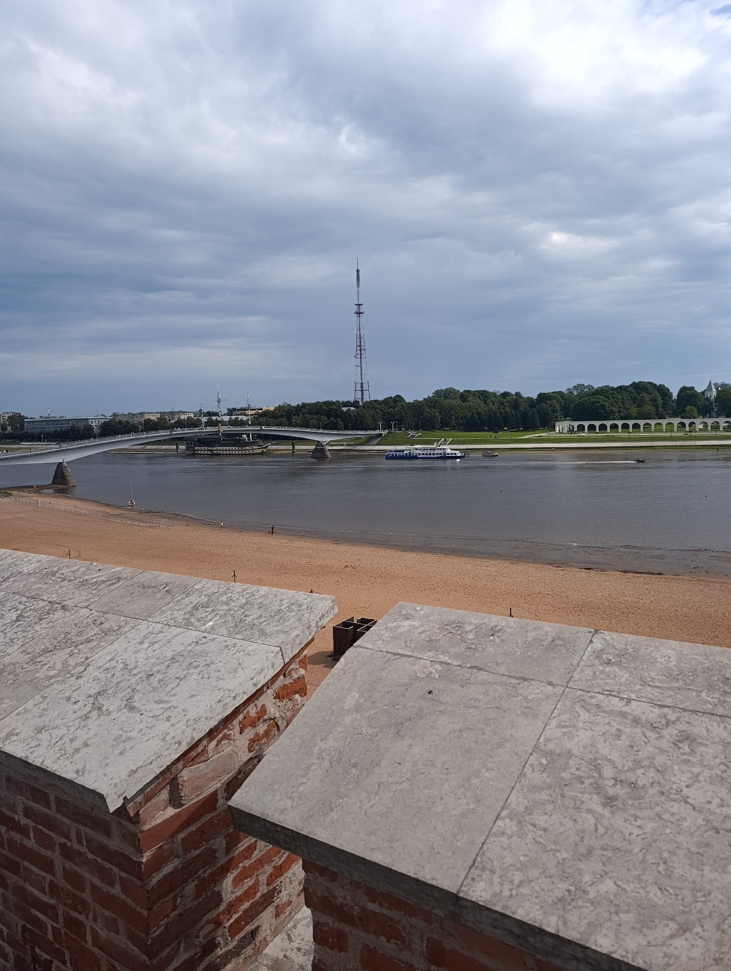 A wide, gray sky with scattered clouds looms over a scene that includes a river, a sandy beach, a bridge, and a distant cityscape with a tall radio tower. In the foreground, a brick wall with a stone cap creates a barrier. The river is calm, with a passenger boat and a smaller boat visible. On the far bank, a line of trees separates the water from buildings and an arcade structure. The beach appears mostly empty, with a few small figures scattered along the shoreline. The overall impression is one of a quiet, somewhat overcast day in an urban setting with natural elements. This scene likely depicts the Volga River embankment in Veliky Novgorod, Russia, given the presence of the distinctive radio tower and the historical architecture.