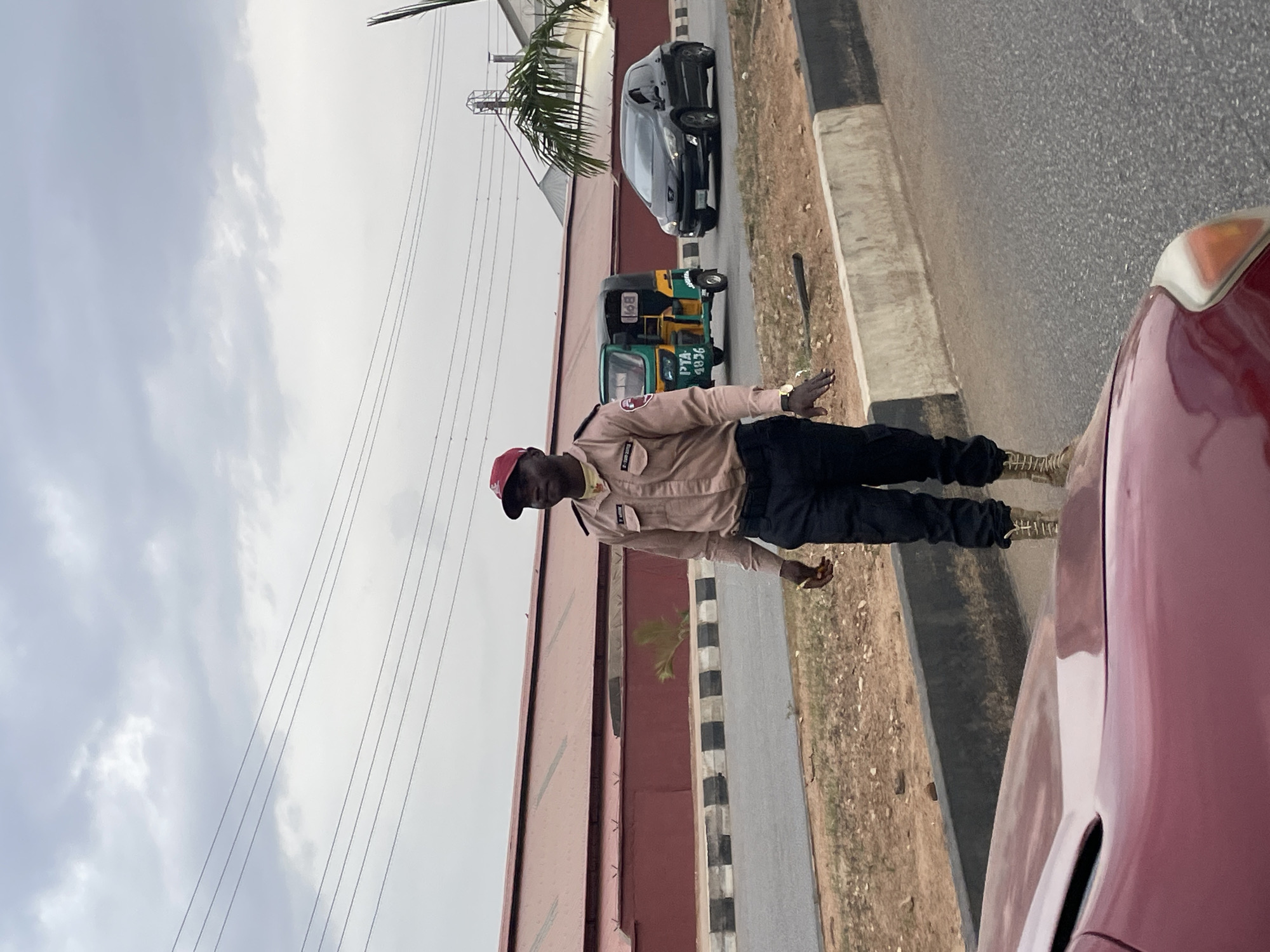 An eye-level shot, rotated 90 degrees counter-clockwise, depicts a man in uniform standing on the side of a road in what appears to be Jos, Nigeria. The man, positioned centrally, wears a light tan or beige long-sleeved uniform shirt with dark trousers and light-colored boots. He has a red cap on his head and a watch on his left wrist. His right arm is outstretched, palm open, as if signaling or directing.
In the background, behind the man, a black sedan is parked or driving on the road, followed by a green and yellow Keke Napep (motorized tricycle) with a passenger. The Keke Napep has "PTA - 4836" clearly visible on its side. Further left, a large building with a reddish-brown facade is seen. The road itself is asphalt, bordered by a concrete curb and dry, rocky ground.
The foreground shows the hood and fender of a red car, suggesting the photograph was taken from inside a vehicle. Overhead power lines are visible against a bright, overcast sky, indicating daytime. The general scene captures a typical street environment in an urban or semi-urban area. The man's expression appears neutral.