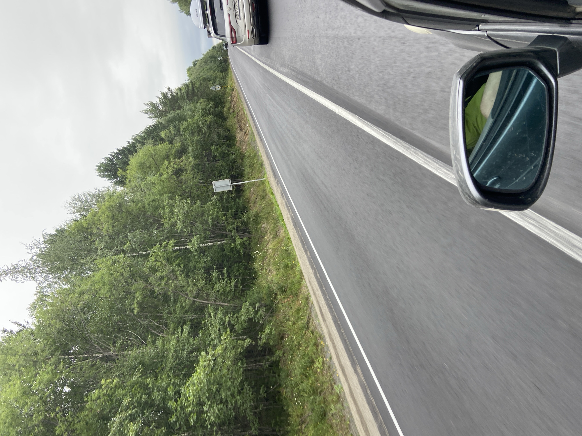 The image depicts a view from a moving vehicle on a paved road bordered by a dense forest on the left and trees on the right. A silver SUV with a roof box and license plate reading "M376TA125" is in the right lane ahead, also moving. The sky is overcast. The road has clear white lane markings. The scene appears to be on a highway or a main road in a rural or semi-rural area. The location is identified as Klyuevka, Russia.