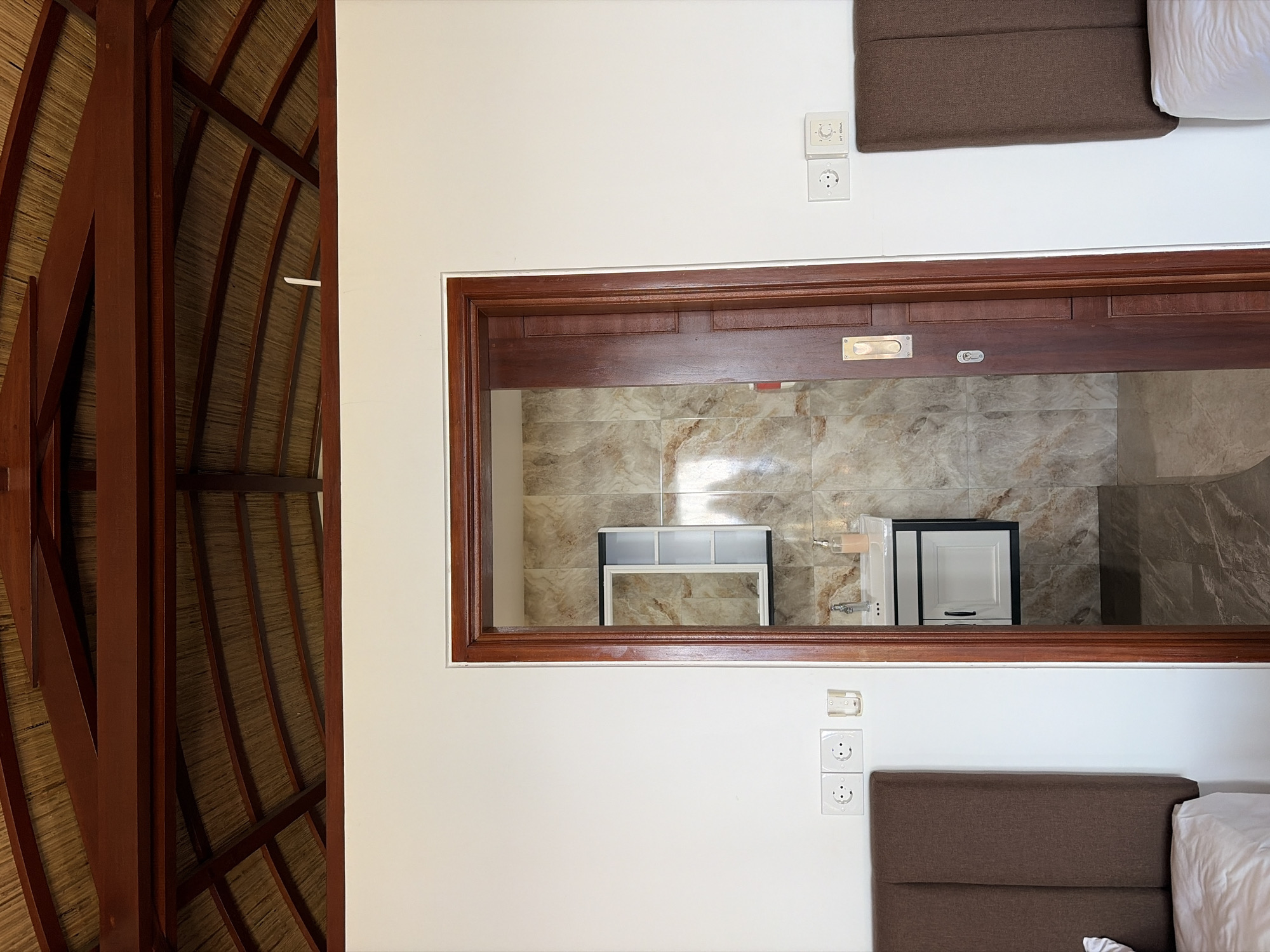 The image depicts the interior of a room with a thatched roof and wooden beams. In the foreground, two beds with brown headboards and white linens are visible on either side of a doorway. The doorway, made of dark wood, leads to a bathroom with tiled walls, a sink, and a cabinet. Electrical outlets and a small glass holder are mounted on the white walls. The scene appears to be in a hotel or guesthouse, likely in Abang, Indonesia, given the architectural style and thatched roof. There are no people or animals visible in the image.