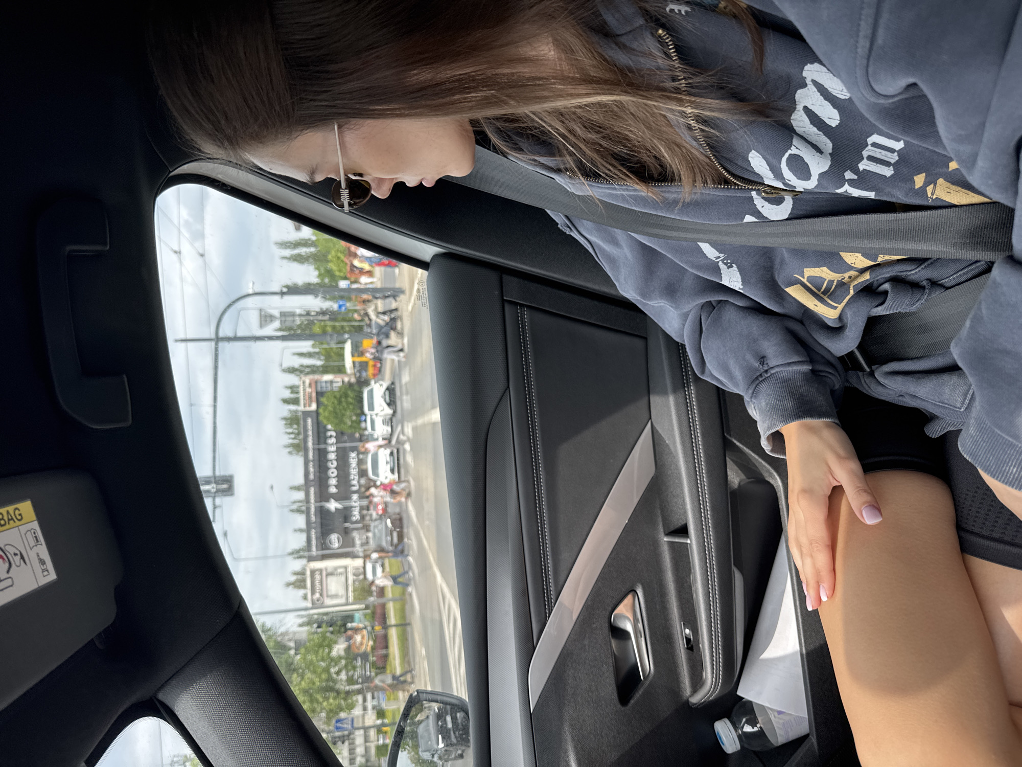 A view from inside a car shows a female passenger wearing sunglasses and a blue hoodie with white and gold text, buckled in with a seatbelt. The car's interior is black with a silver accent on the door. Outside the car window, a city street scene includes buildings, cars, streetlights, and pedestrians. A sign reading "PROGRESJA SALON LAZIENEK" is visible on a building. A water bottle is in the door pocket near the passenger's leg. The woman's hand rests on her upper leg. The location is in Unknown, Unknown.