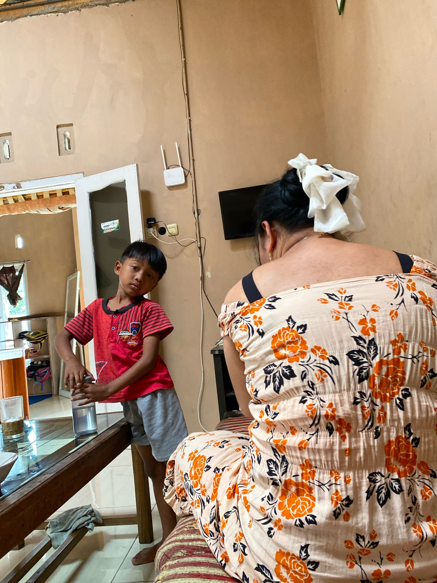 A young boy stands by a wooden table in what appears to be a living room or dining area of a home in Pesantren, Indonesia. The boy is wearing a red and black striped t-shirt with graphics and gray shorts. He is holding a clear water bottle. To his right, a woman is seated, viewed from the back, wearing a light-colored dress with a floral pattern of orange flowers and dark leaves. She has her dark hair tied up with a white cloth. Behind the boy, a white door with a glass panel is slightly ajar, revealing a glimpse of another room with shelves containing items. A Wi-Fi router is mounted on the wall above a television. The walls of the room are a neutral, light brown color, and the floor appears to be tiled. A glass of liquid and other items are on the table in front of the boy. The lighting suggests it is daytime, possibly indoors with some natural light.