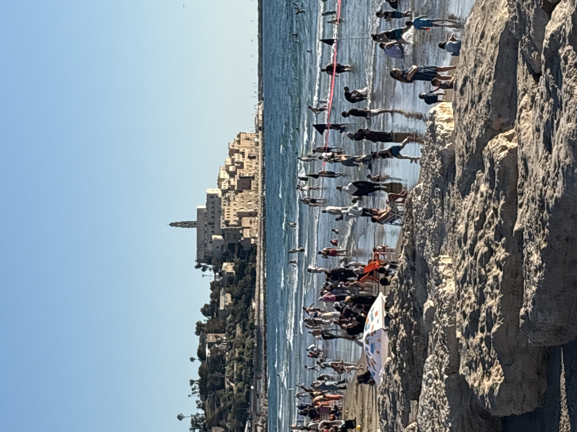 The image depicts a busy beach scene in Tel Aviv, Israel, likely on a sunny day. The foreground is dominated by large, textured rocks, suggesting a natural coastline. Beyond the rocks, a crowd of people are gathered on the sand and in the shallow water. Many are standing, some appear to be participating in an organized activity, possibly a race or competition, indicated by red and black flags and lines in the water. The water is a vibrant blue and appears somewhat choppy with small waves. In the background, a densely built city with light-colored buildings rises from a green, hilly landscape. A prominent tower is visible, which could be a landmark. The sky is a clear, bright blue, reinforcing the impression of good weather. The atmosphere appears lively and active, with many people enjoying the beach. There is no visible text in the image.
