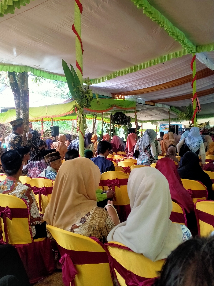 Under a decorated canopy, many people are seated in rows of yellow chairs with red ribbons, facing forward. Most of them are wearing headscarves in shades of white, beige, red, and black. Some wear traditional hats. Green and red fabric is wrapped around support poles, and greenery is added to the decor. A few trees are visible outside the tent-like structure.