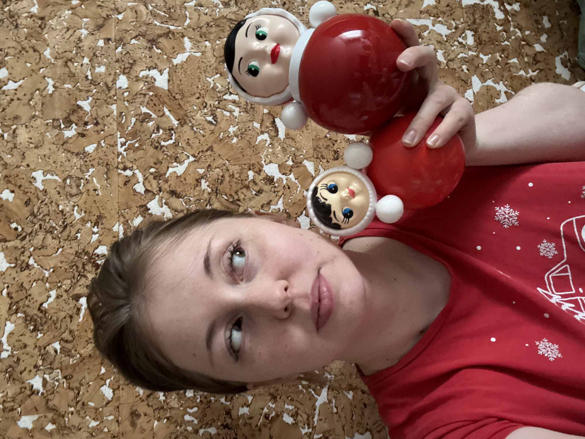 A woman holds two roly-poly dolls against a backdrop of what appears to be cork flooring or wall covering. The woman is fair-skinned and wearing a red top with a snowflake design. She is looking upward to the left, with a slightly pensive expression. The dolls are traditional in style, with round red bodies and stylized painted faces. The location is Unknown, Unknown.