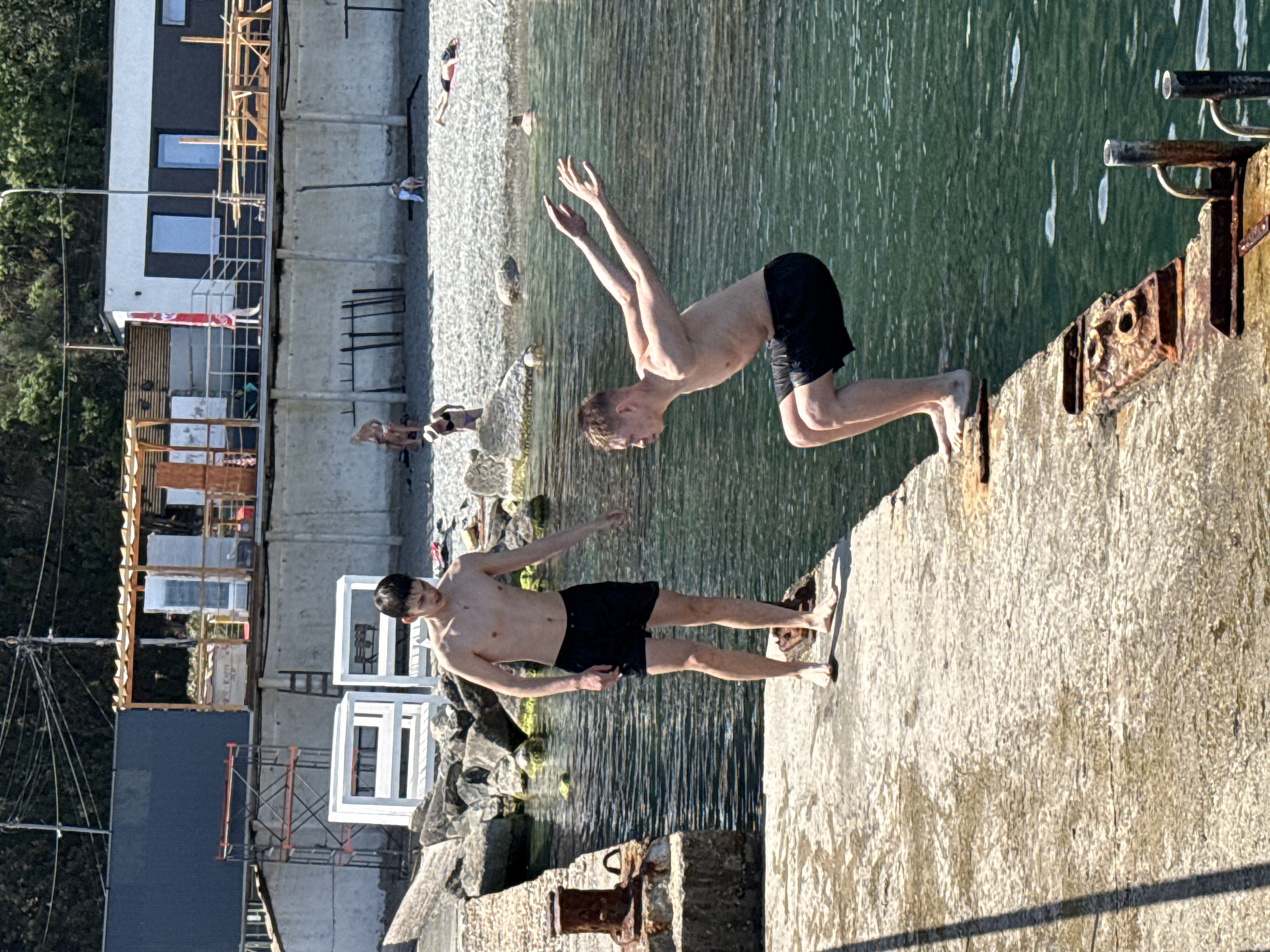 Two shirtless young men are captured on a sunlit concrete dock, one poised mid-air, arms raised, ready to dive into the green water. The second figure stands on the dock's edge, facing left, ready to jump. In the background, a beach stretches along the shoreline, with various people and structures including buildings, scaffolding, and concrete walls visible. The location is Unknown, Unknown.