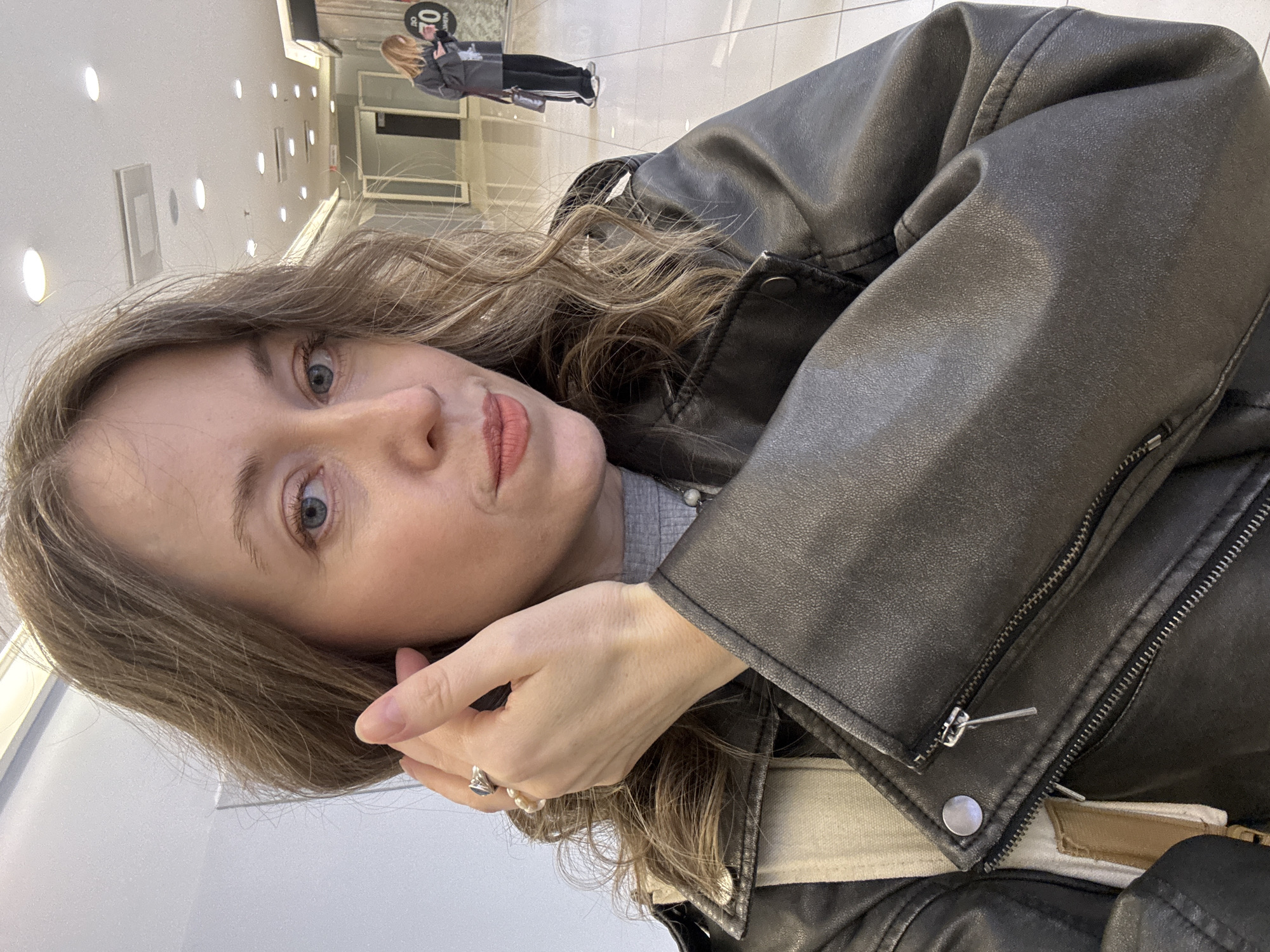 The image shows a woman in a black leather jacket indoors. She is fair-skinned with light brown hair and blue eyes. She is wearing a ring on her finger. In the background, a person is visible in a hallway lined with recessed lights. The scene appears to be inside a shopping mall or large building. There is insufficient information to identify the location in Saint Petersburg, Russia.