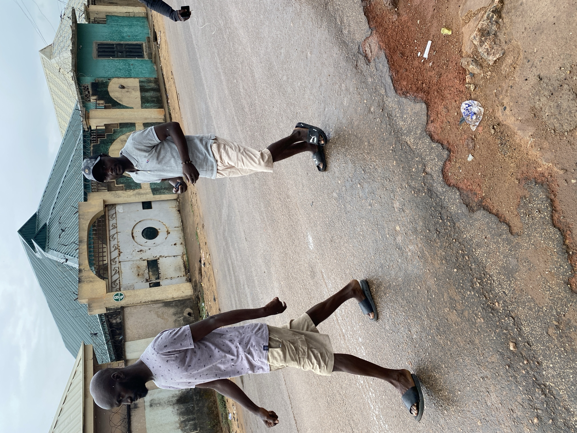 Two dark-skinned men are standing on a paved road in Narayi, Nigeria. The man on the left is bald, wearing a lavender shirt with white dots and beige shorts, and black sandals. The man on the right is wearing a grey shirt, beige shorts, a black baseball cap, and black sandals. Behind them, there's a building with a corrugated metal roof, faded blue paint, and a metal gate. The road surface is cracked and damaged, with dirt and debris along the edges. A person's hand holding a phone is visible in the upper left corner. The sky is overcast.