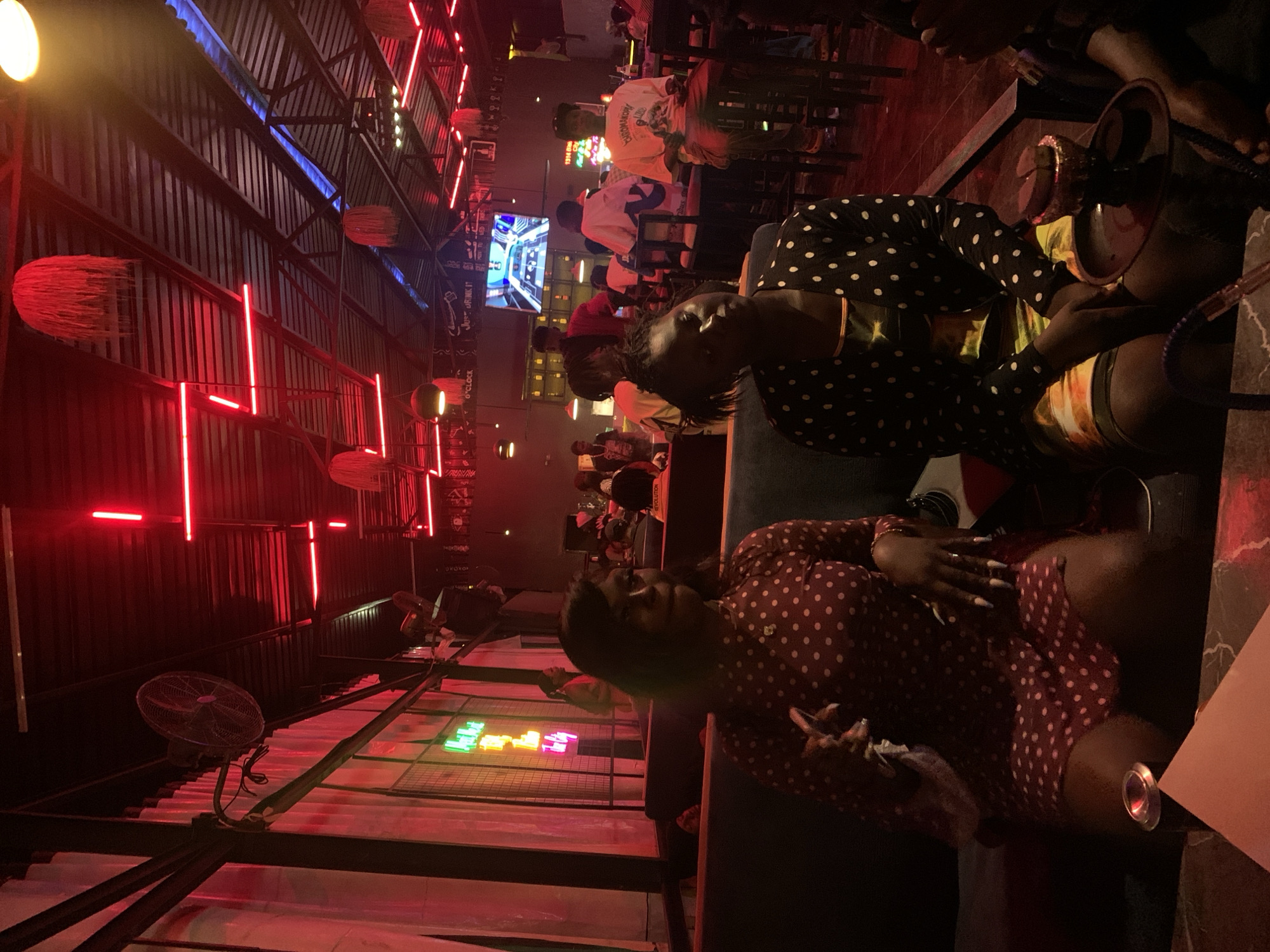 The image depicts a dimly lit, indoor establishment on Rukuba Road, Nigeria, characterized by vibrant red neon lighting and a dark, corrugated metal ceiling. Two women are seated on a dark-colored couch in the foreground. The woman on the left wears a dark red dress with white polka dots and has long, light-colored fingernails. She is holding a white object in her right hand. The woman on the right wears a black and white polka dot cardigan over a yellow and orange patterned top, and she is resting her hands on her lap. A hookah is visible on a small table to the right of the woman on the right. In the background, other patrons are seated at tables and on couches, and a large screen television displays a video game. The overall atmosphere appears to be social and relaxed, possibly a lounge or nightclub.