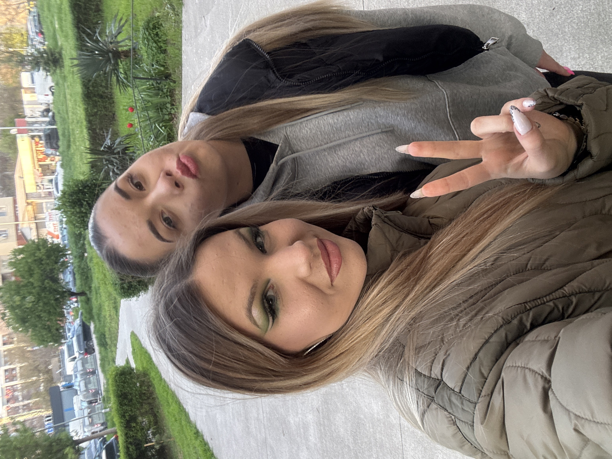 Two young women are posing closely for a selfie outdoors. The woman in the foreground wears a tan quilted jacket and light-brown hair with blonde highlights. She has green eye makeup. The other woman is wearing a gray hoodie with a black vest. The background includes buildings, green spaces with shrubbery, trees, and parked cars, indicating an urban environment in Plovdiv, Bulgaria. The woman in the foreground is displaying a peace sign with her hand.