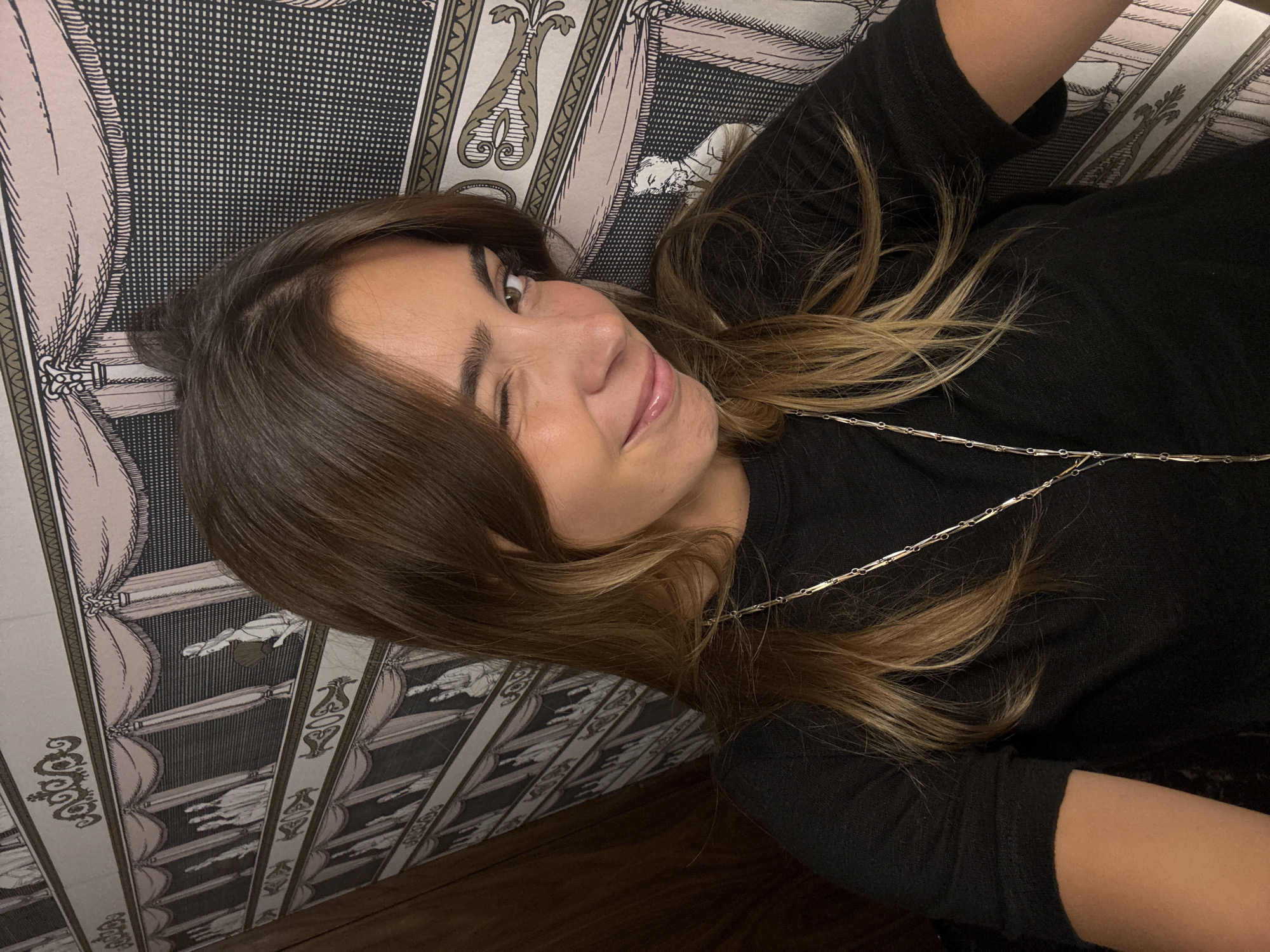 A young woman is lying down, winking, in Vila Verde, Portugal. She has medium-length brown hair with blonde highlights, a necklace and is wearing a black top. She is lying on a patterned surface. The pattern consists of grey and pink stripes. Within these stripes are black designs and what looks like drawings of people. A wooden floor is visible.