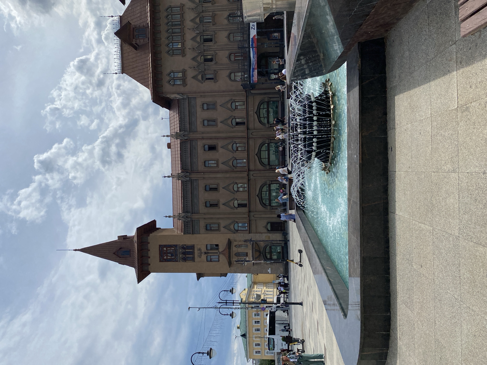 In the city of Unknown, Unknown, a fountain is situated in a paved plaza in front of a large, ornate building with a reddish-brown tiled roof. The building has several stories, with numerous windows of varying sizes and architectural styles. A prominent tower with a pointed spire rises from one section of the building.
The fountain, in the foreground, is filled with clear blue water that is actively spraying upwards, creating a dynamic visual. The fountain's basin is lined with dark, polished stone.
Several people are visible in the scene. In the distance, some individuals are walking across the plaza, while others appear to be interacting near the building's entrance. A woman in green pants and a white jacket stands near the fountain, facing left. Several scooters are parked or being ridden by people in the background.
The sky above is filled with a mix of white, puffy clouds and darker, more ominous clouds, suggesting a partly cloudy day. Streetlights with decorative elements are also visible in the mid-ground. In the far background, other buildings and a white bus can be seen.