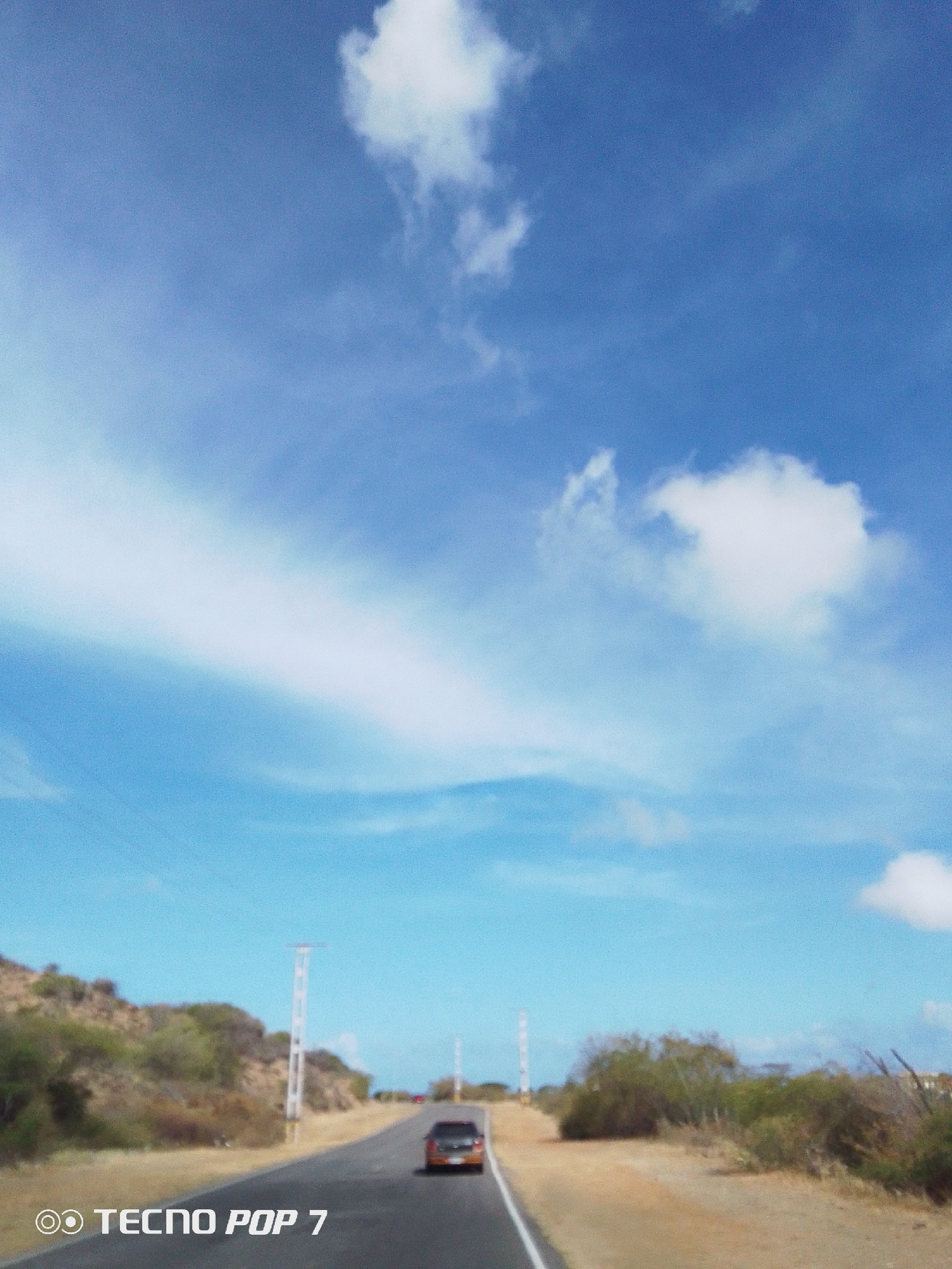 The image depicts a road in what appears to be a rural or semi-arid environment. The sky is predominantly blue with scattered white clouds. On either side of the road, there are patches of low-lying vegetation, possibly bushes and shrubs. A car is visible on the road, moving away from the camera. Tall, white structures resembling poles or towers stand along the roadside at intervals. The image includes a "Tecno POP 7" watermark. The city is identified as Pedro González, Venezuela.