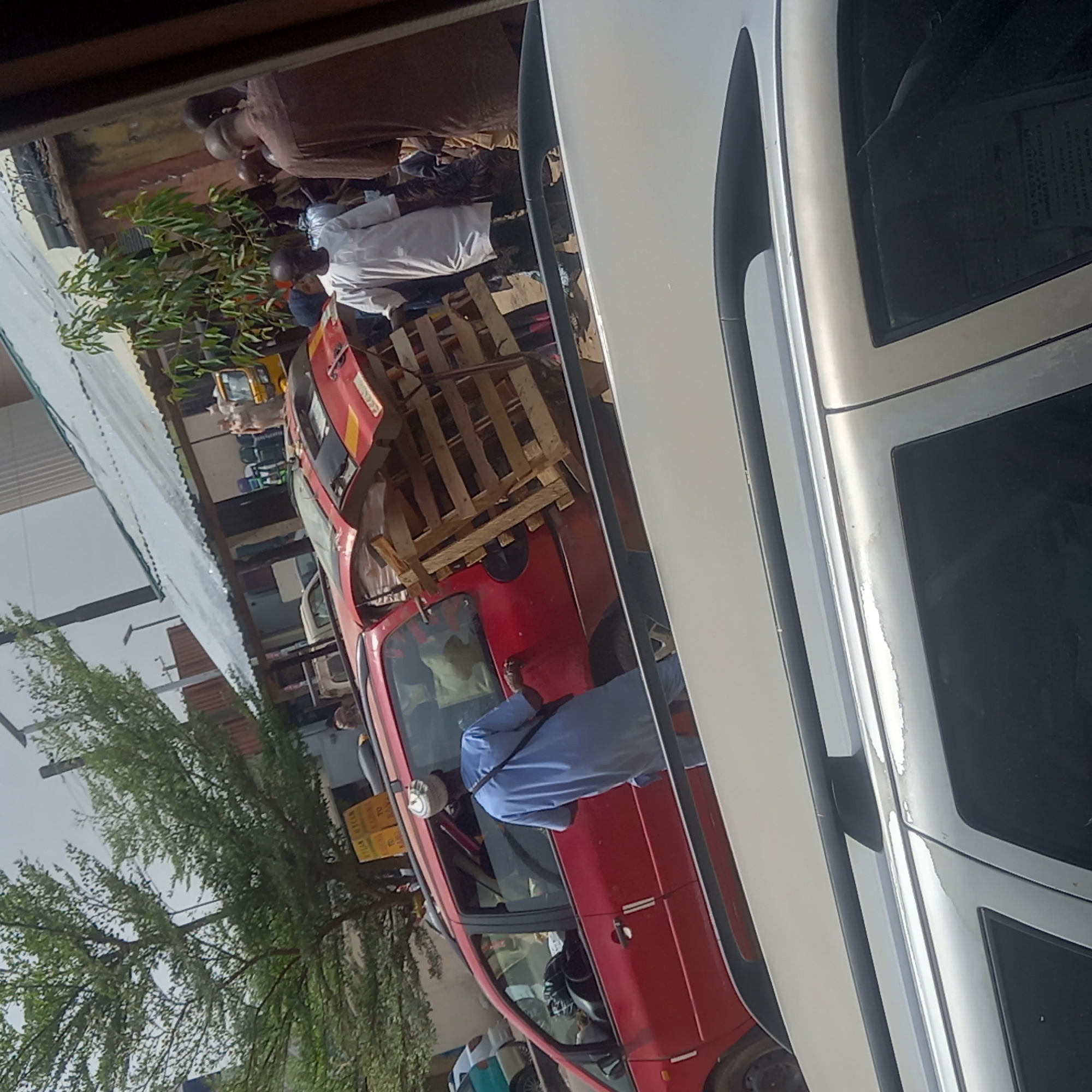 The image depicts a street scene in Dadi Riba, Nigeria, with a red car overloaded with wooden pallets secured on its roof. People are standing around the car. A white car is partially visible in the foreground. The buildings and trees suggest an urban environment.