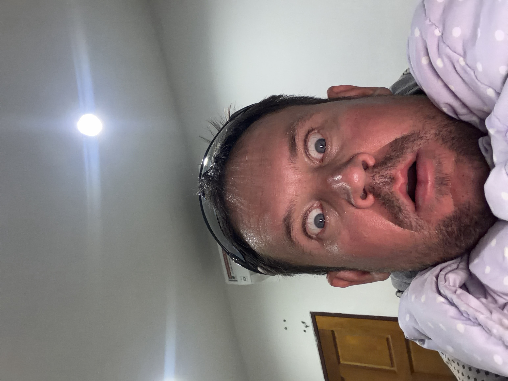 In Baan Mae Nam, Thailand, a selfie is captured from a low angle looking up at a man's face. The man has a surprised expression with wide eyes and an open mouth. He is wearing sunglasses perched on his forehead, and his face is flushed and appears to be sweating. He is wrapped in a light purple blanket with white polka dots. Behind him, the wall is white, and a light fixture is visible on the ceiling. To the left, a wooden door is partially visible. An air conditioning unit is mounted on the wall above and behind the man's head. There are also some small, dark objects on the wall to the left of the air conditioning unit.