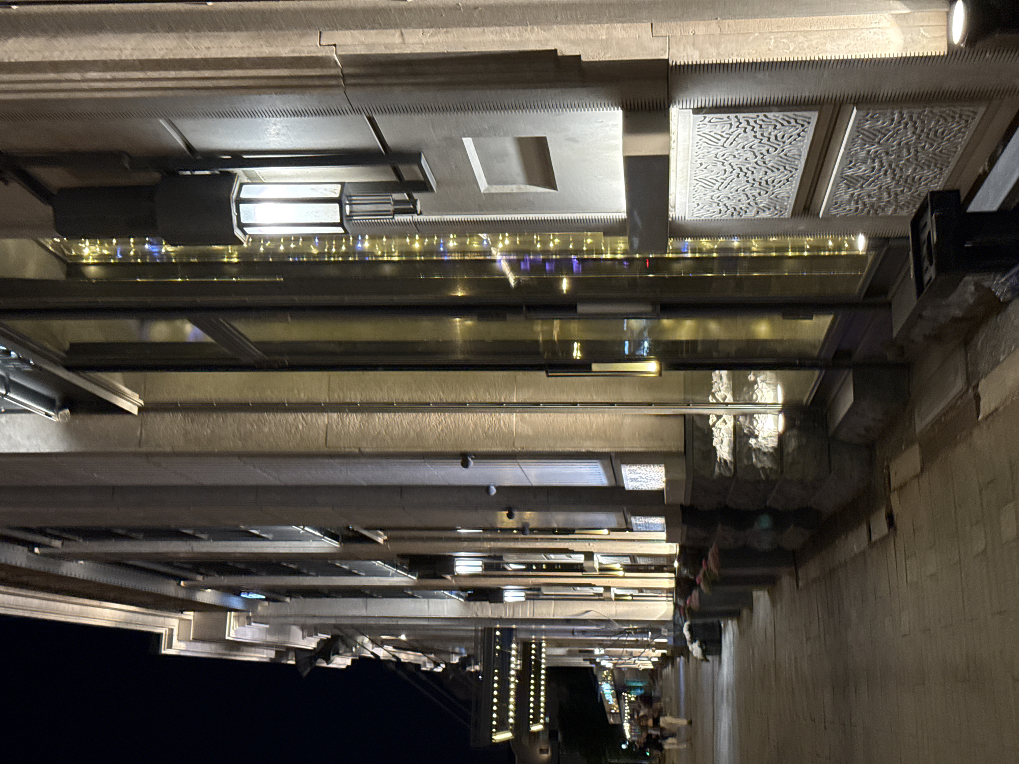 The image displays a night scene along an illuminated urban street in Moscow, Russia. The prominent subject is the well-lit facade of a multi-story building, characterized by light-colored stone or concrete. This building features prominent vertical pilasters, horizontal cornices, and numerous rectangular windows. The ground floor incorporates large display windows, through which interior lighting and decorative strands of warm white and blue fairy lights are visible.
The building's facade is extensively illuminated by upward and downward-pointing spotlights, highlighting its architectural details. Wall-mounted lanterns with frosted glass panels are spaced along the facade, with one positioned prominently in the foreground on the right. The lower sections of the facade are adorned with decorative panels featuring complex, organic, or abstract relief patterns, and some pilasters exhibit vertical striations. A rectangular indentation is also visible on one of the wall sections.
To the left of the building is a wide, paved pedestrian sidewalk constructed from rectangular pavers, extending into the background. A series of dark, cylindrical planters containing flowers or foliage are arranged along the building's edge. Several indistinct human figures are seen walking along the sidewalk in the mid-ground, indicating pedestrian activity. Further down the street, an extended awning or canopy, adorned with multiple parallel rows of small, bright white lights, stretches over a section of the sidewalk. In the immediate foreground, on the right side of the sidewalk, a dark, upright stand is partially visible. The sky is dark, consistent with a nocturnal setting. The architectural style, extensive illumination, and urban elegance are characteristic of central Moscow's renovated historic and commercial districts, such as Tverskaya Street or Kamergersky Lane.